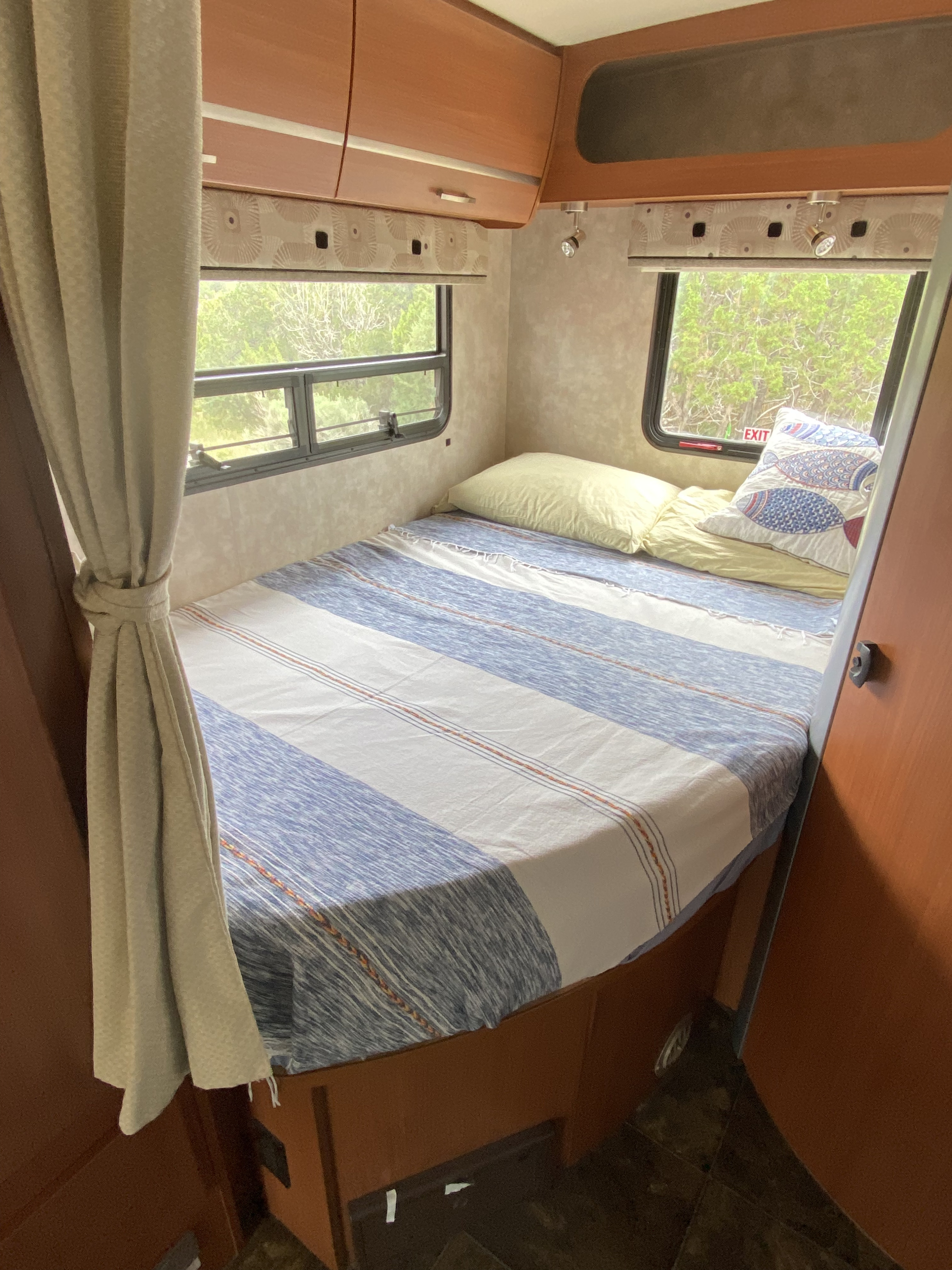 RV rental bedroom features a comfy striped bed, yellow pillows, overhead storage, and nature views.. Winnebago View 2013