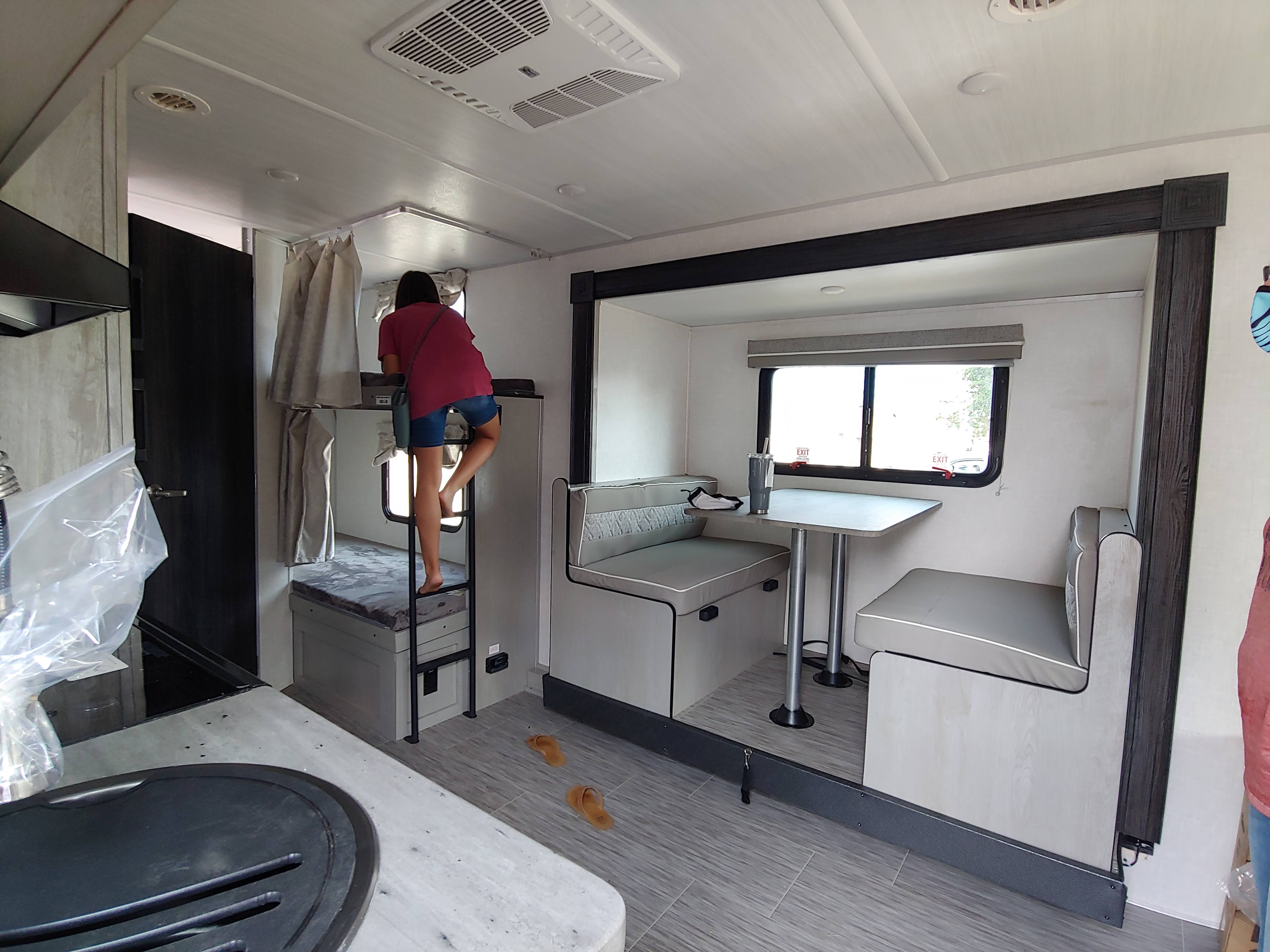 Inside a modern RV rental: a woman climbs bunk beds. Features include a dinette, kitchen area, and ample living space.. Forest River Ibex 2021