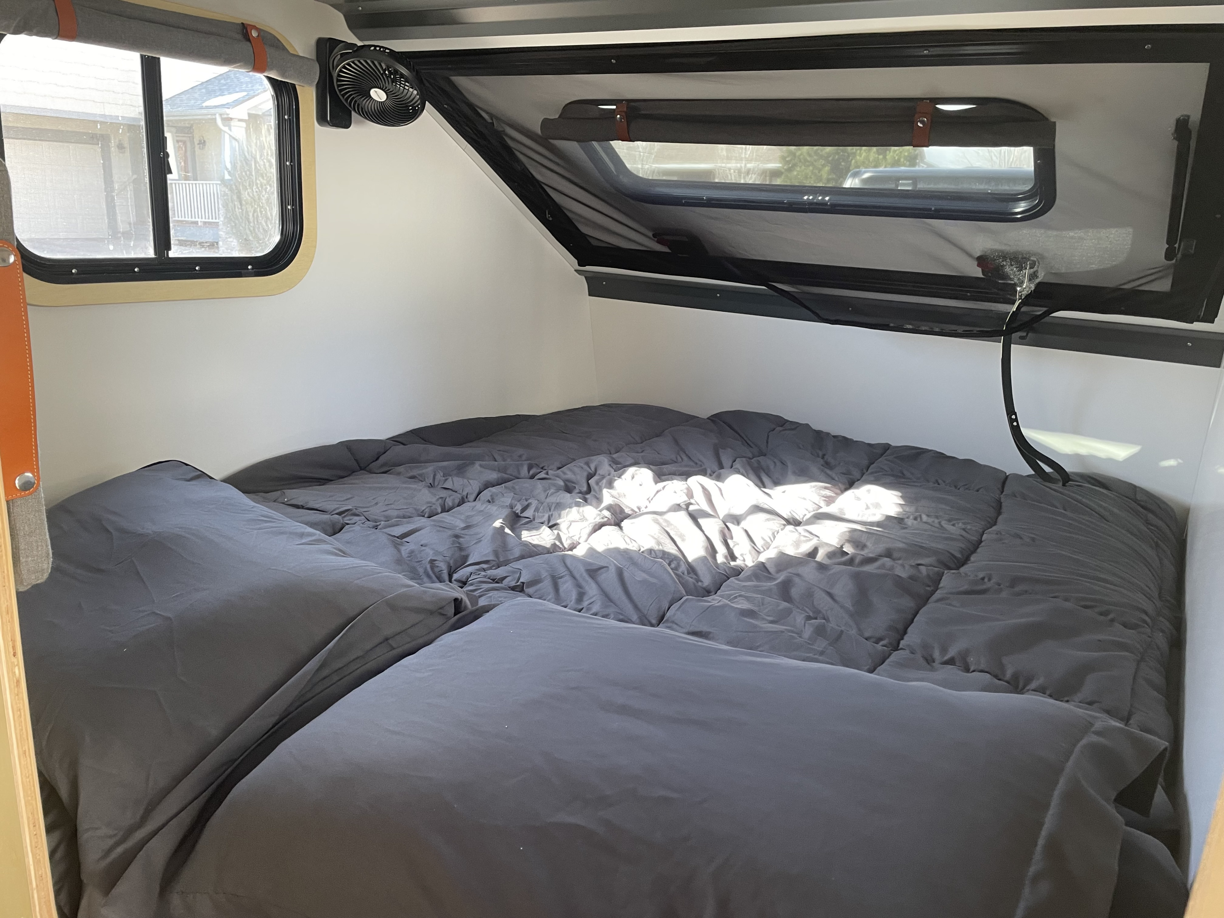 View inside an rv rental: a large grey bed with sunlit bedding, windows, and a fan, creating a cozy sleeping space.. campinawe sport trailer 2023