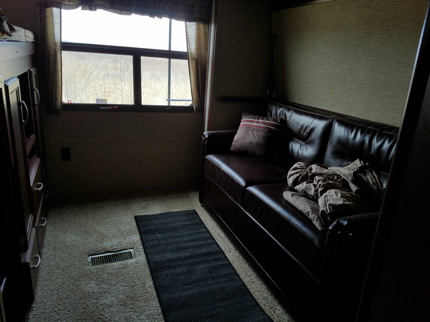 Inside an RV rental, a leather sofa sits opposite a window and wood storage unit with bunk above. Comfortable travel living.. Keystone Outback 2016