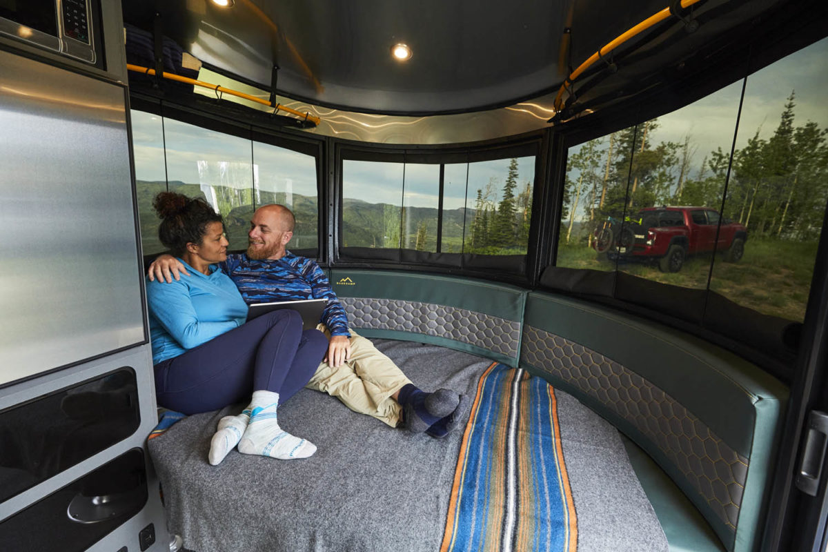 Couple relaxes inside a cozy RV rental, enjoying panoramic views of mountains, forests, and a truck with a bike. Ideal for nature escapes.. Airstream Basecamp 2022
