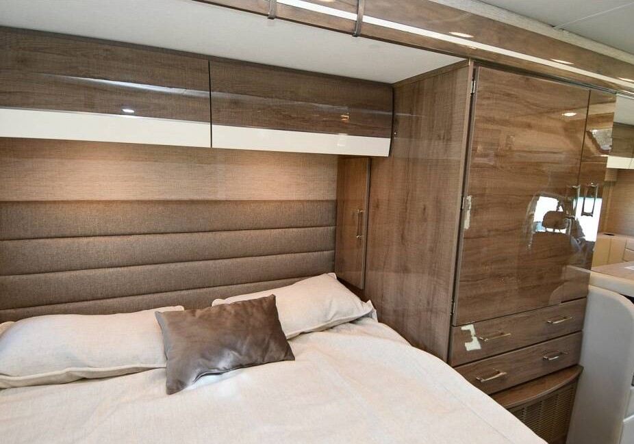 Spacious RV rental bedroom featuring a large bed, padded headboard, sleek overhead cabinets, and a tall wardrobe with drawers.. Jayco Melbourne 2023