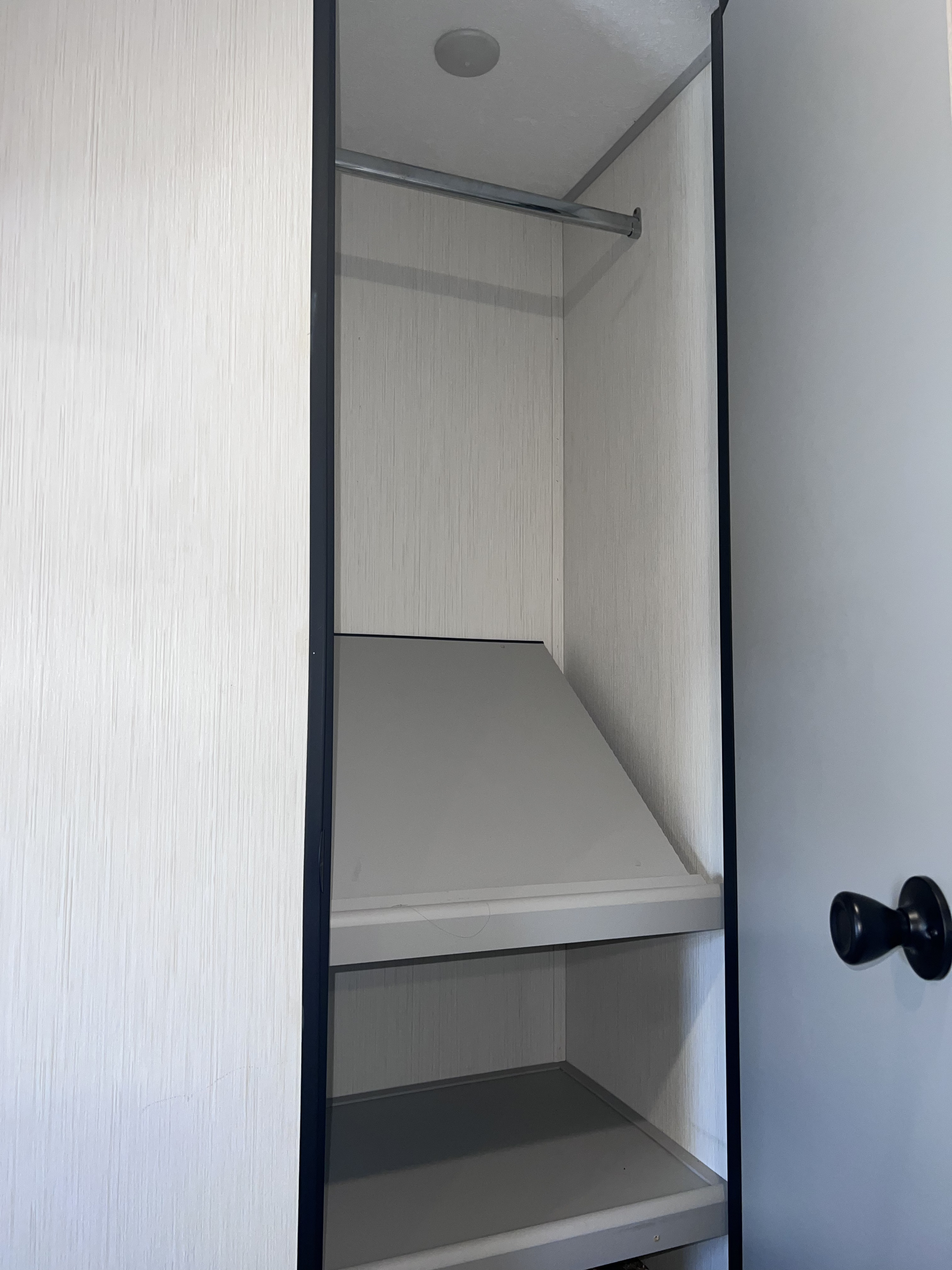 Empty closet in an RV rental, featuring a top hanging rod, angled shelf, and lower flat shelf, with an interior light.. Forest River Vibe 2021