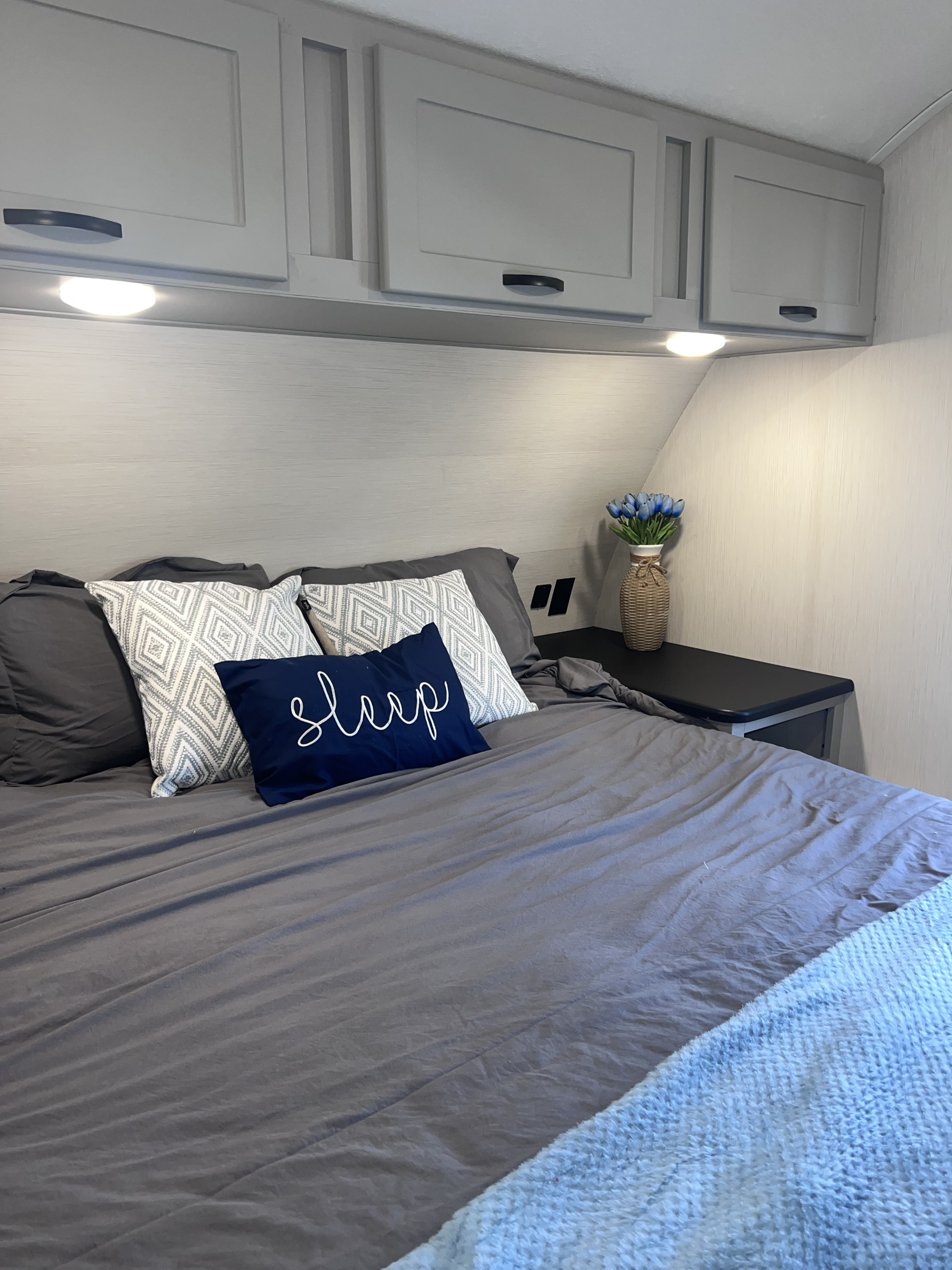 Inviting rv rental bedroom: gray bed with patterned/sleep pillows, overhead cabinets, under-cabinet lights, and blue flowers.. Forest River Vibe 2021