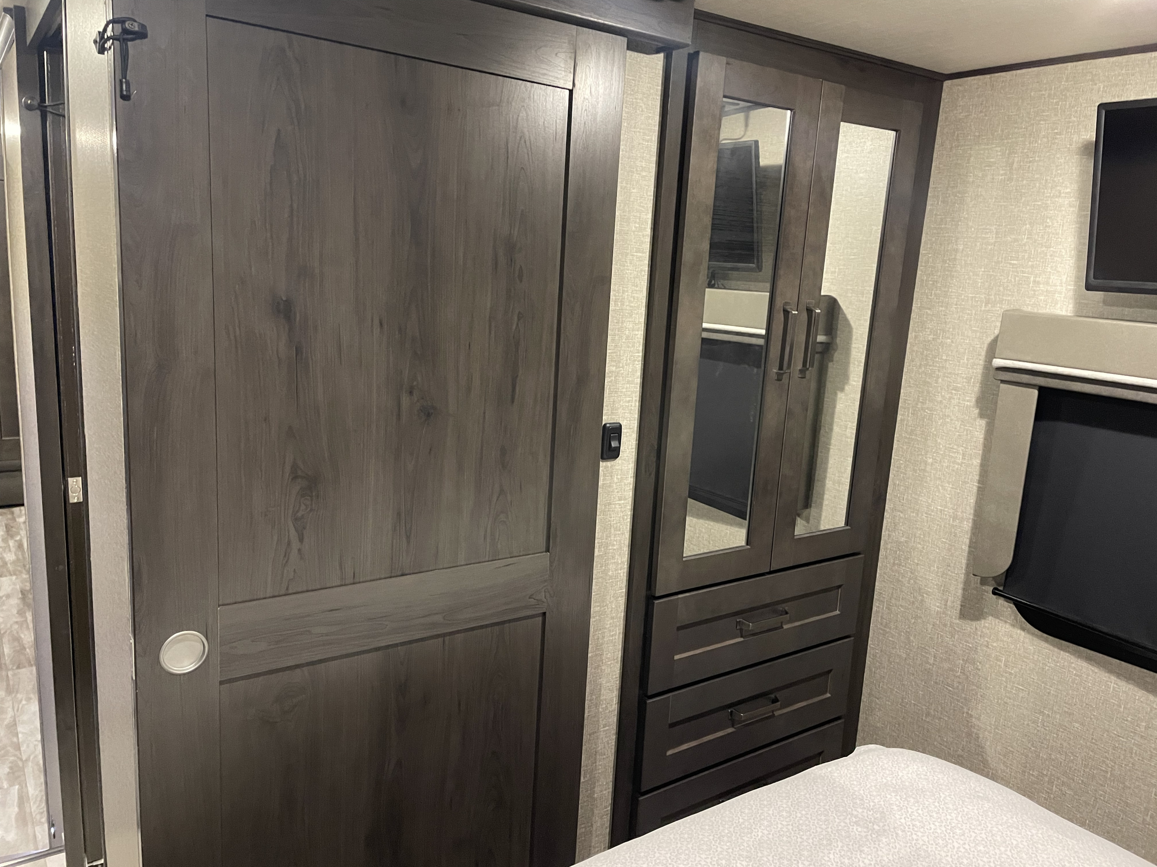 RV rental bedroom: dark wood sliding door, mirrored wardrobe with drawers, wall-mounted TV, and window shade, offering compact comfort.. Grand Design Reflection 2021