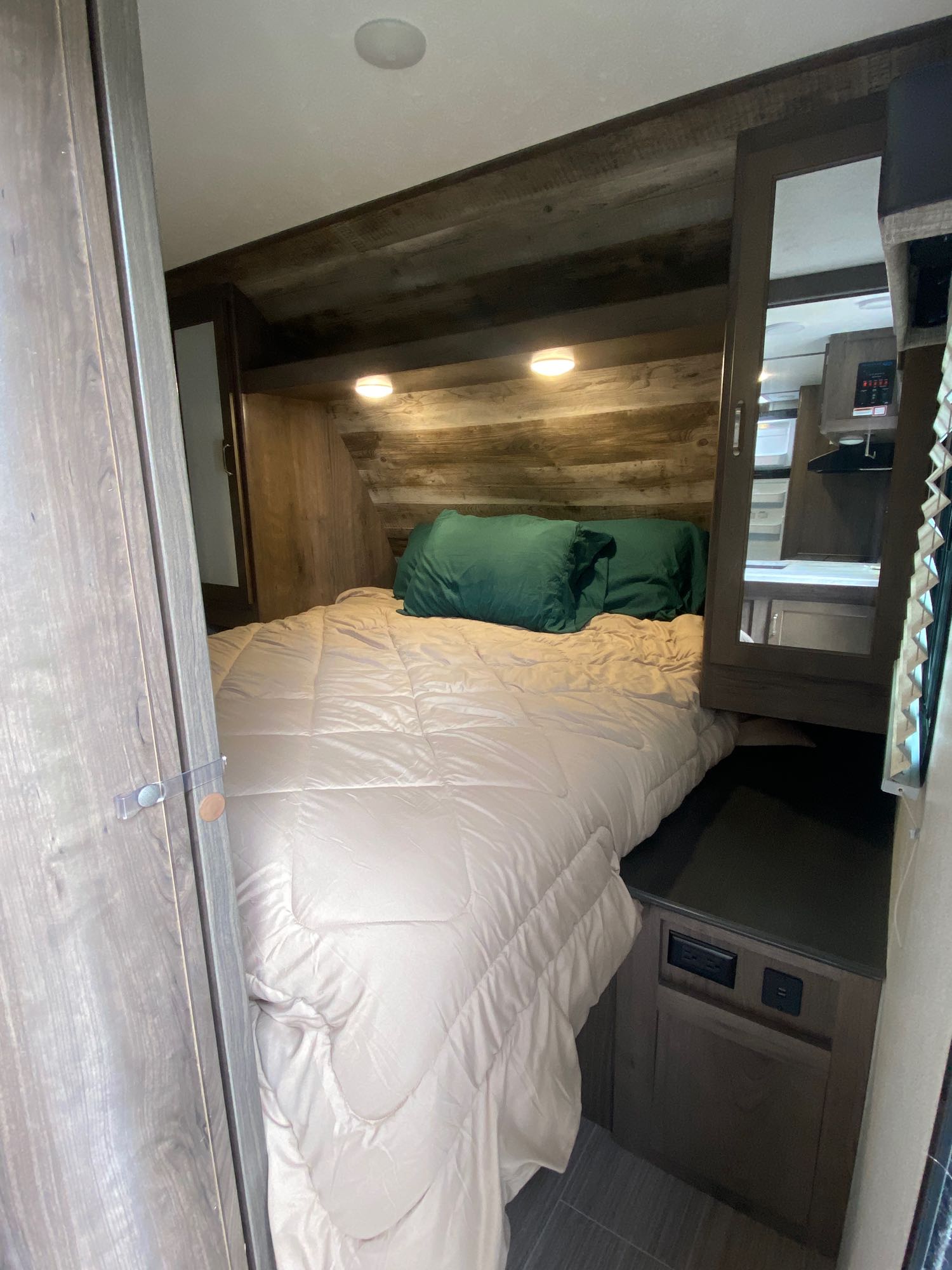 Cozy RV rental bedroom showcasing a full bed, built-in storage, and charging ports. Mirrored cabinet & wood paneling complete the compact space.. Keystone RV Passport SL 2021
