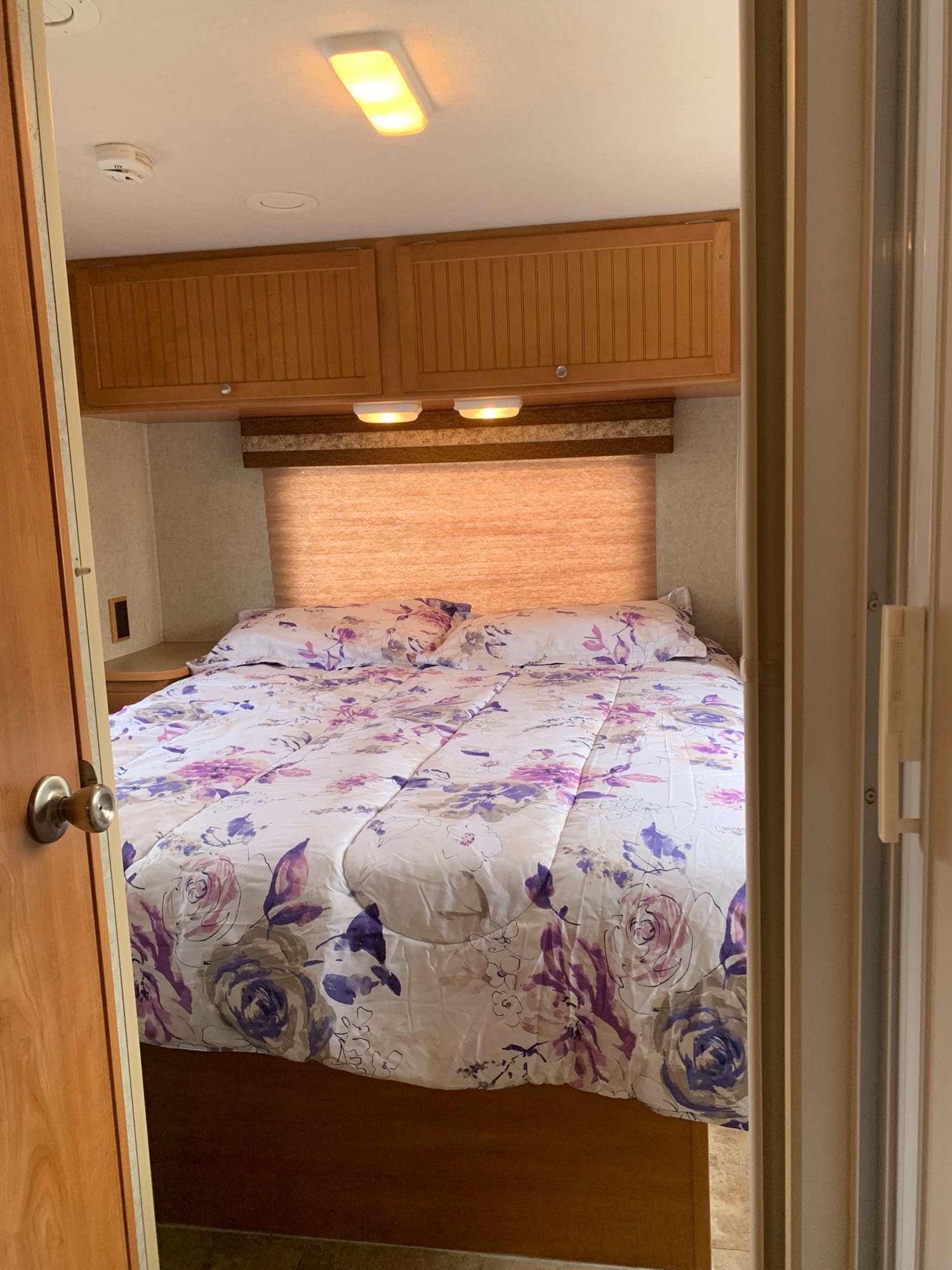 Step into this cozy RV rental bedroom. Features floral bed, wood cabinets, smoke detector, and warm lighting.. Winnebago Access 2007