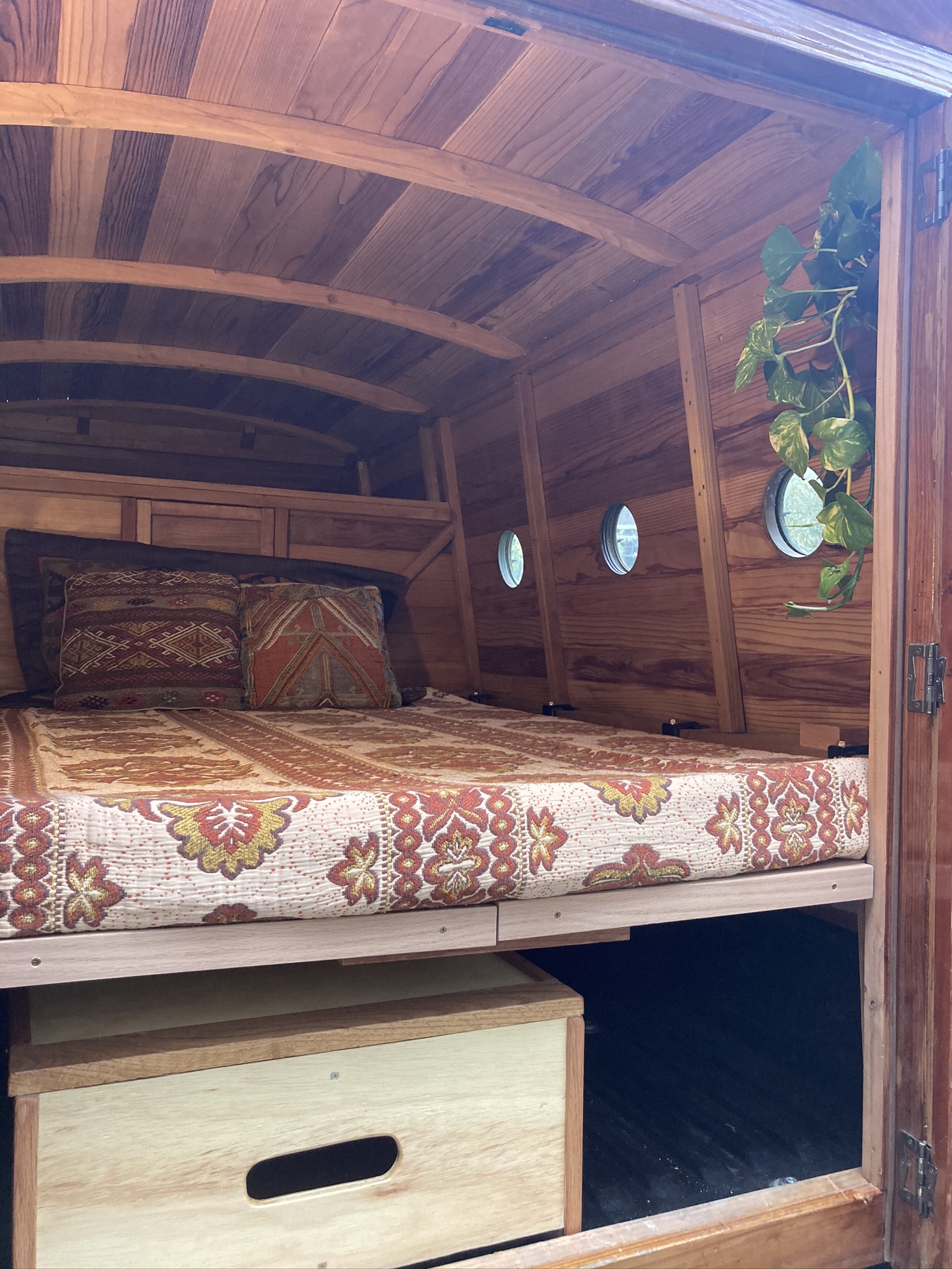 Rustic RV rental interior: wood-paneled walls & ceiling, cozy bed with patterned linens, porthole windows, and a plant.. Custom Camper 2017