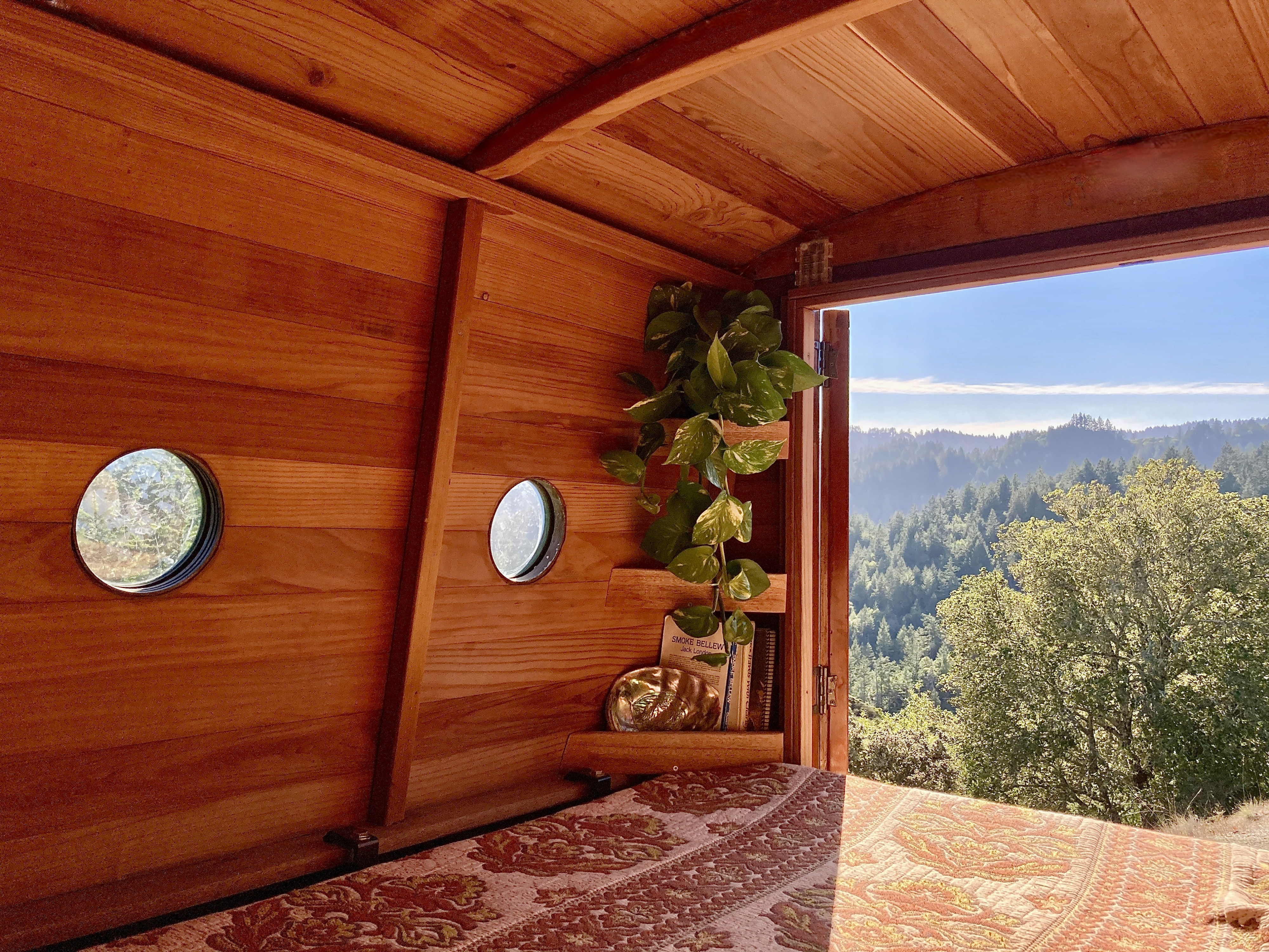Cozy wooden interior of an **rv rental** features porthole windows, a bed, and an open door to a panoramic forest view under a blue sky.. Custom Camper 2017