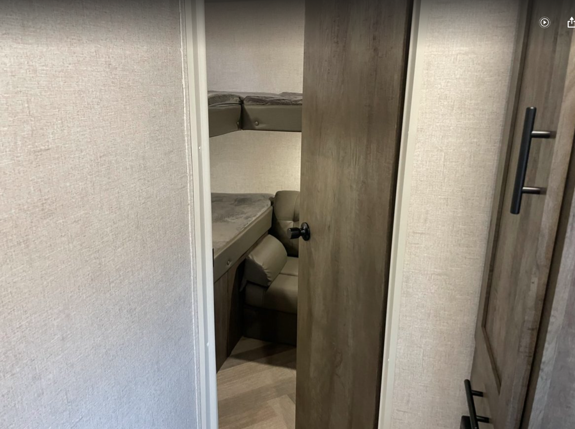 RV rental interior displaying a compact bunk room with two beds and a cozy seating area. A wooden door is visible to the right.. Forest River Wildwood X-Lite 2021
