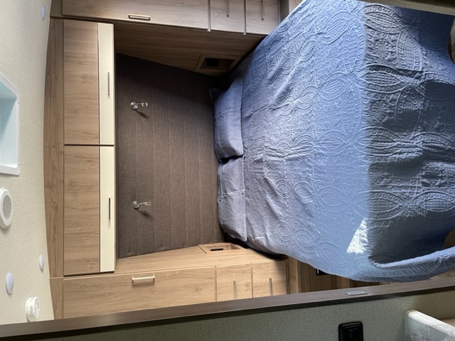 Relax in this RV rental bedroom: comfy bed with a blue quilt, practical wood cabinets & drawers.. Grand Design Imagine 2022