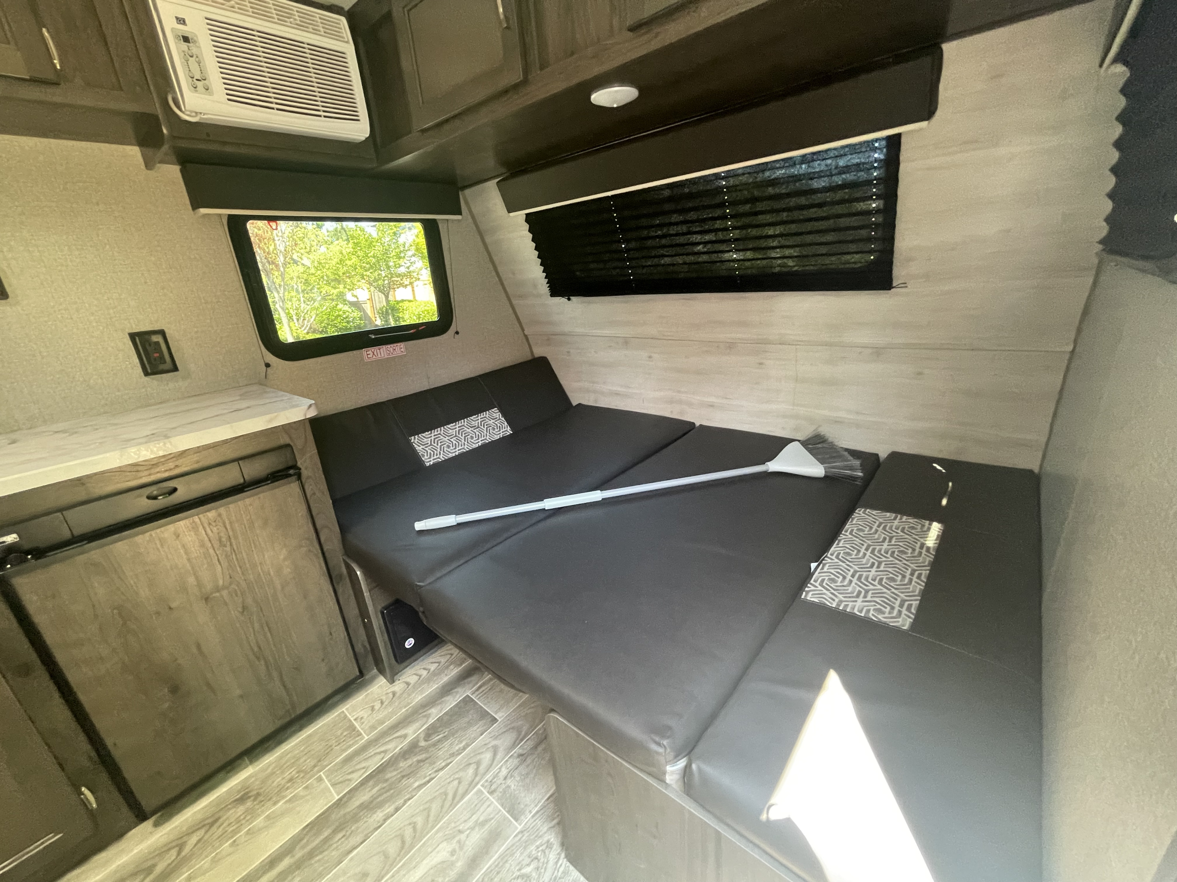 Compact rv rental interior showing a dark grey fold-out bed, window with blind, AC unit, and built-in fridge.. Jayco Jay Flight 2021