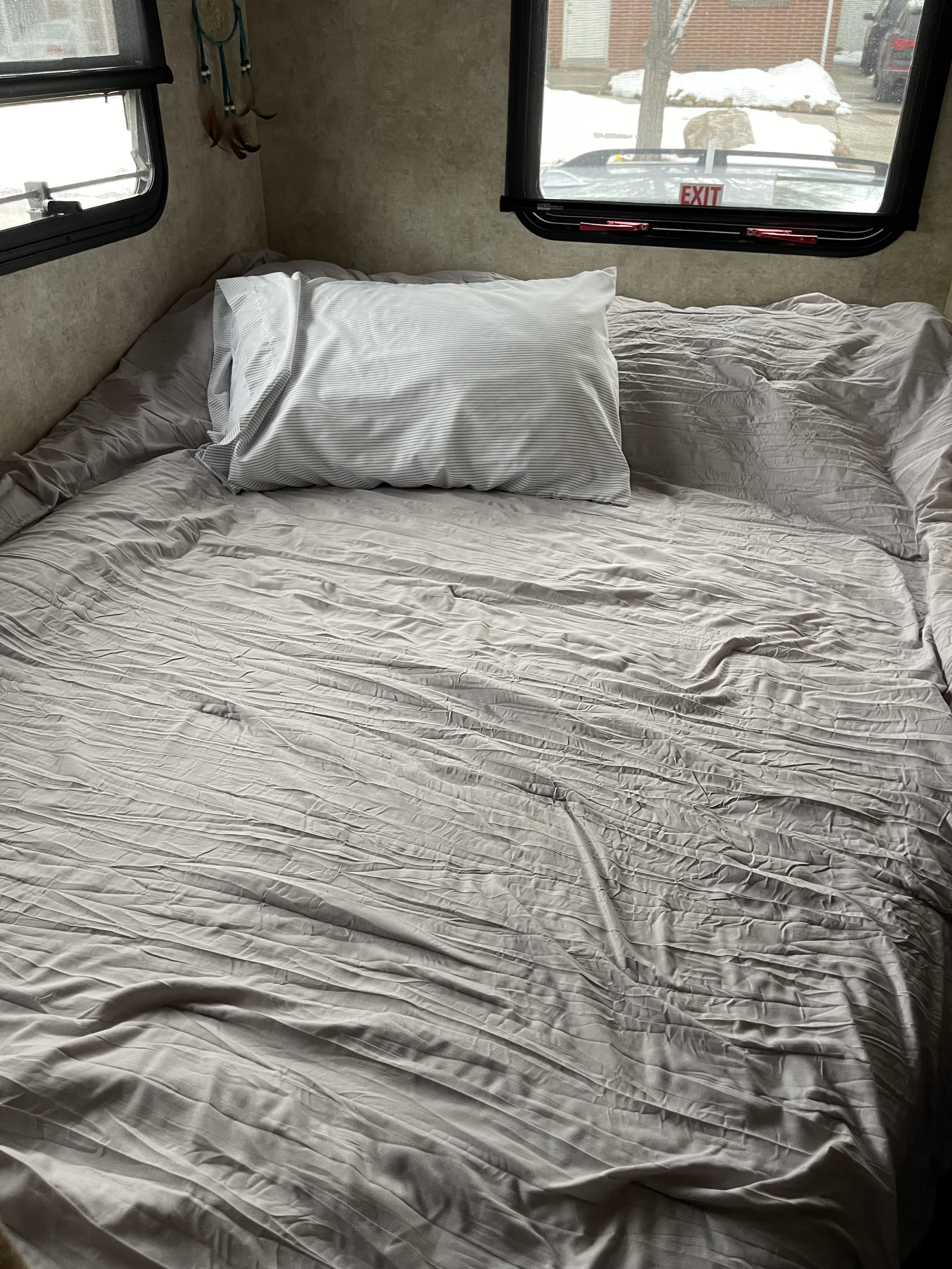 RV rental bedroom: A gray, wrinkled bed with striped pillow. Windows show snowy outdoors and an "EXIT" sign.. Winnebago View 2011