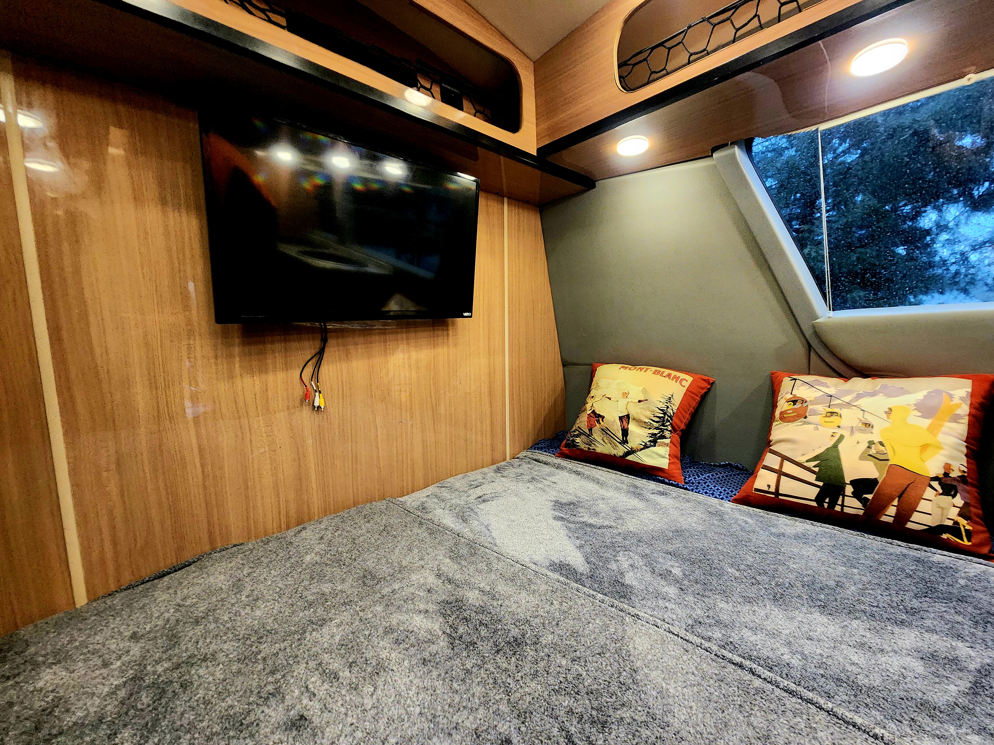 RV rental interior: cozy bedroom with a comfy bed, wall-mounted TV, and ski-themed pillows. A window shows trees outside.. Other Other 2016