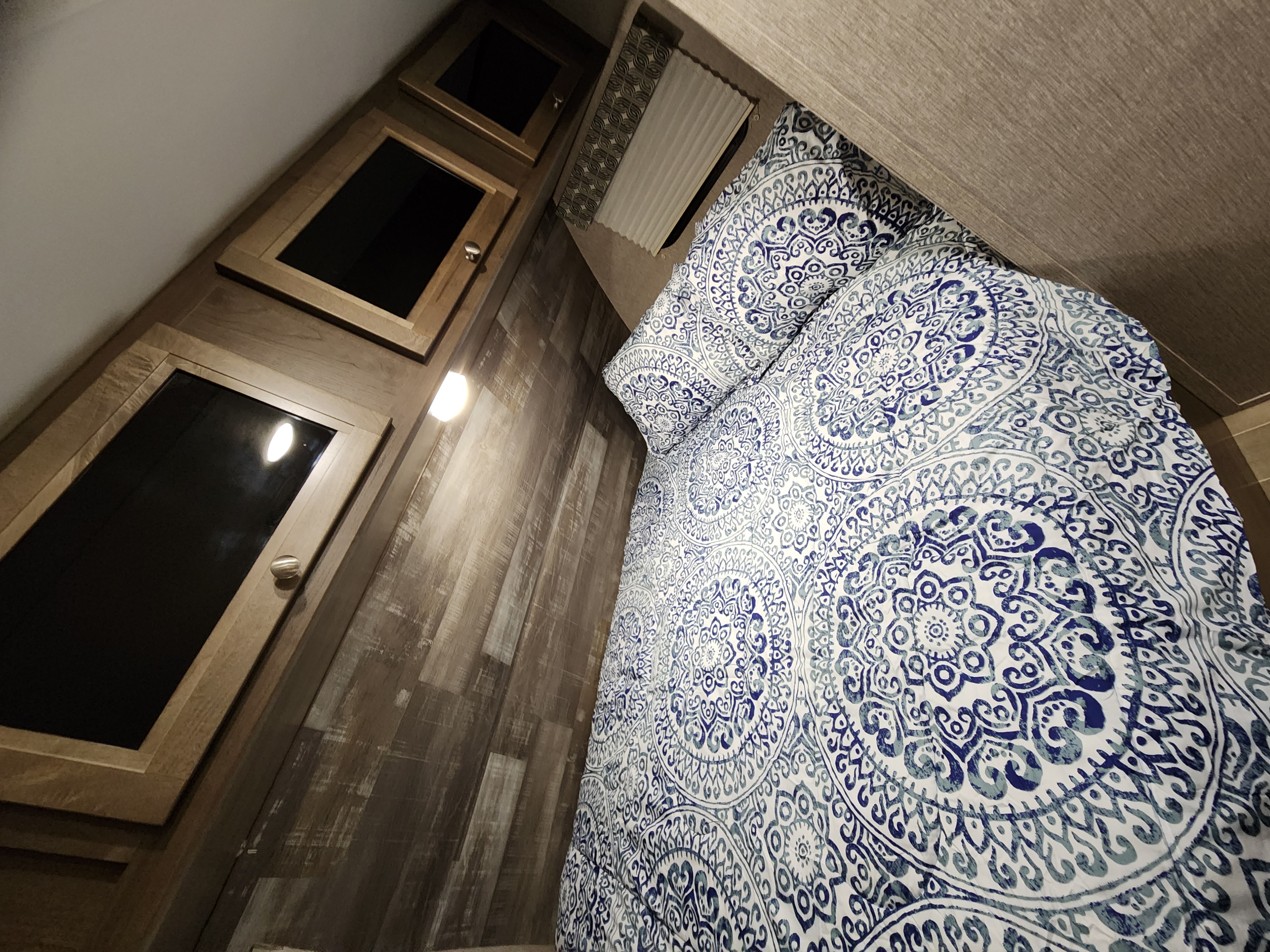 An RV rental's cozy bedroom, featuring blue mandala patterned bedding, wood cabinets, and paneling.. Dutchmen Coleman Light LX 2020