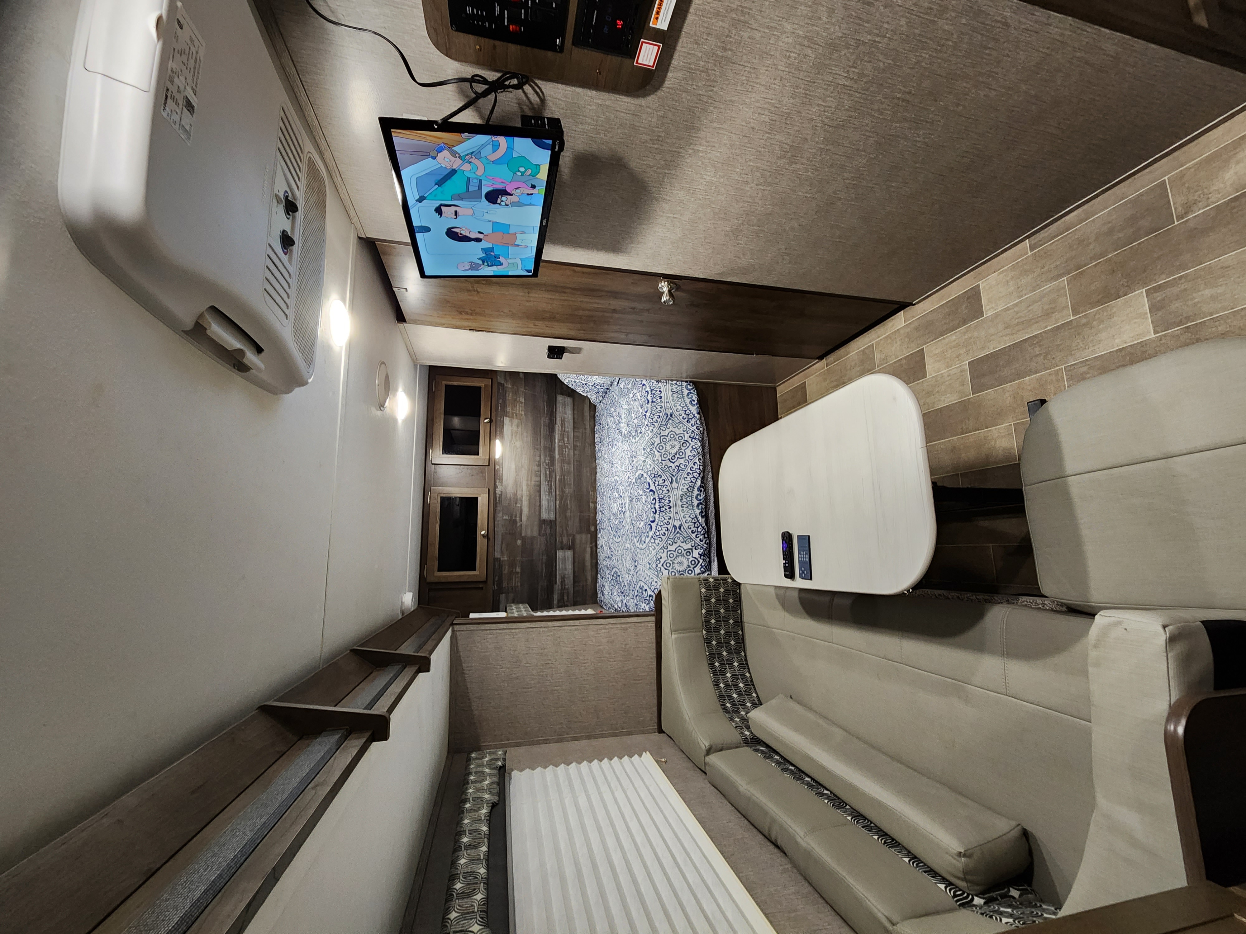 Interior view of an RV rental showing a compact bedroom, dining area, and TV playing a cartoon.. Dutchmen Coleman Light LX 2020