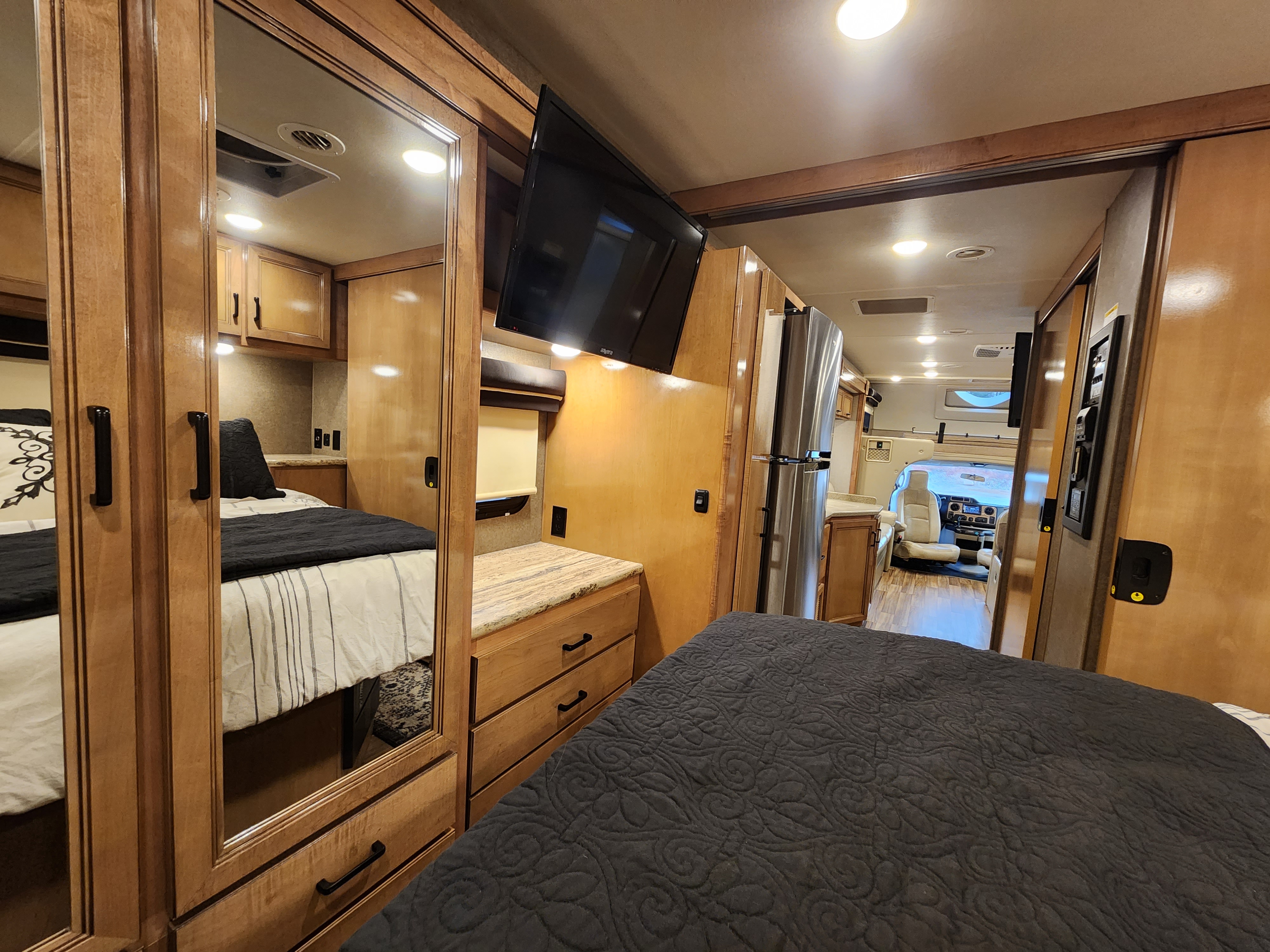 Spacious RV rental interior: bedroom, mirrored wardrobe, TV, fridge, and front cabin. Warm wood cabinetry & bright lights.. Thor Motor Coach Quantum 2019