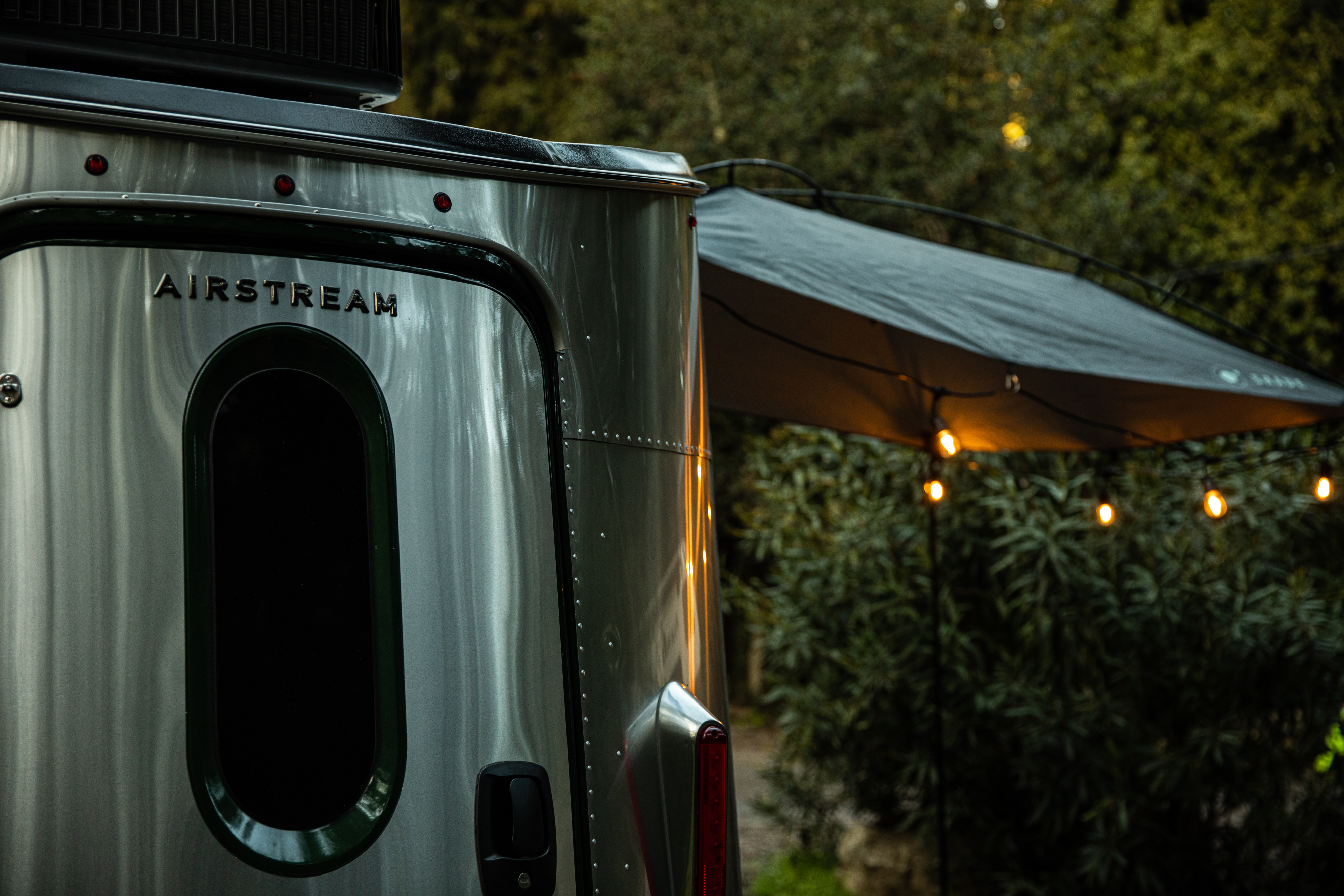 An iconic silver Airstream RV rental parked outdoors at dusk, with its distinctive logo, an extended awning, and warm string lights.. Airstream Other 2023