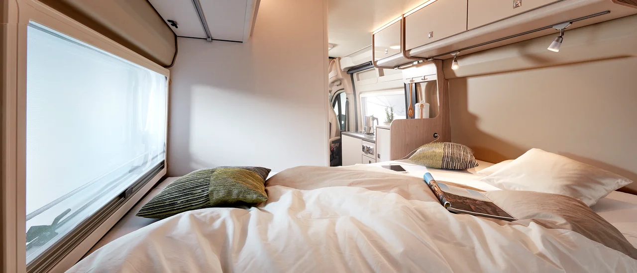 Cozy RV rental interior: bedroom with comfy bed, large window, and connected compact kitchen. Perfect for travel.. Bürstner Campeo C600 Toit relevable Black Forest 2023