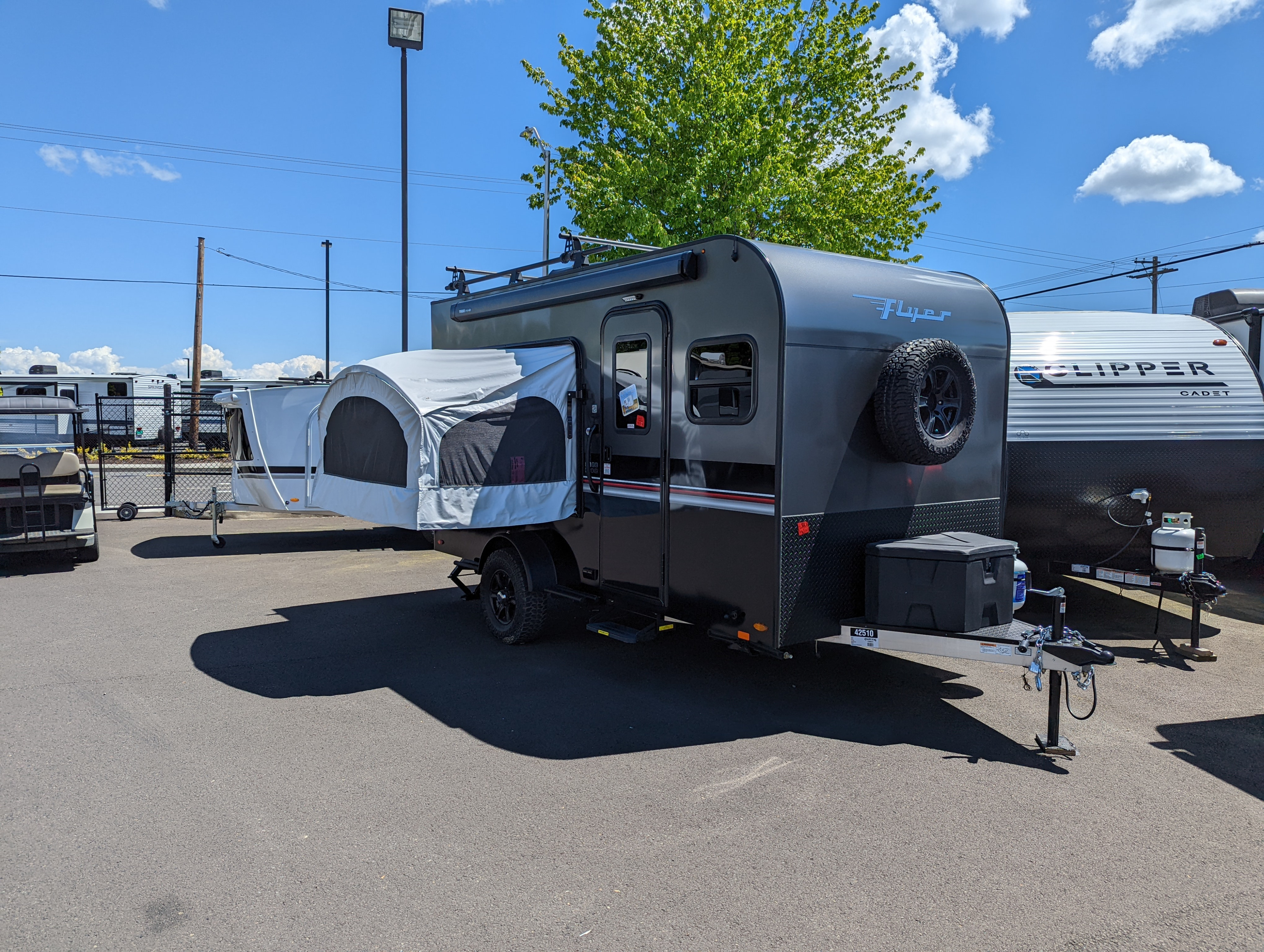 A dark grey "Flyer" off-road RV rental trailer featuring a side pop-out tent. A white "Clipper" camper is behind it.. InTech RV Flyer 2022