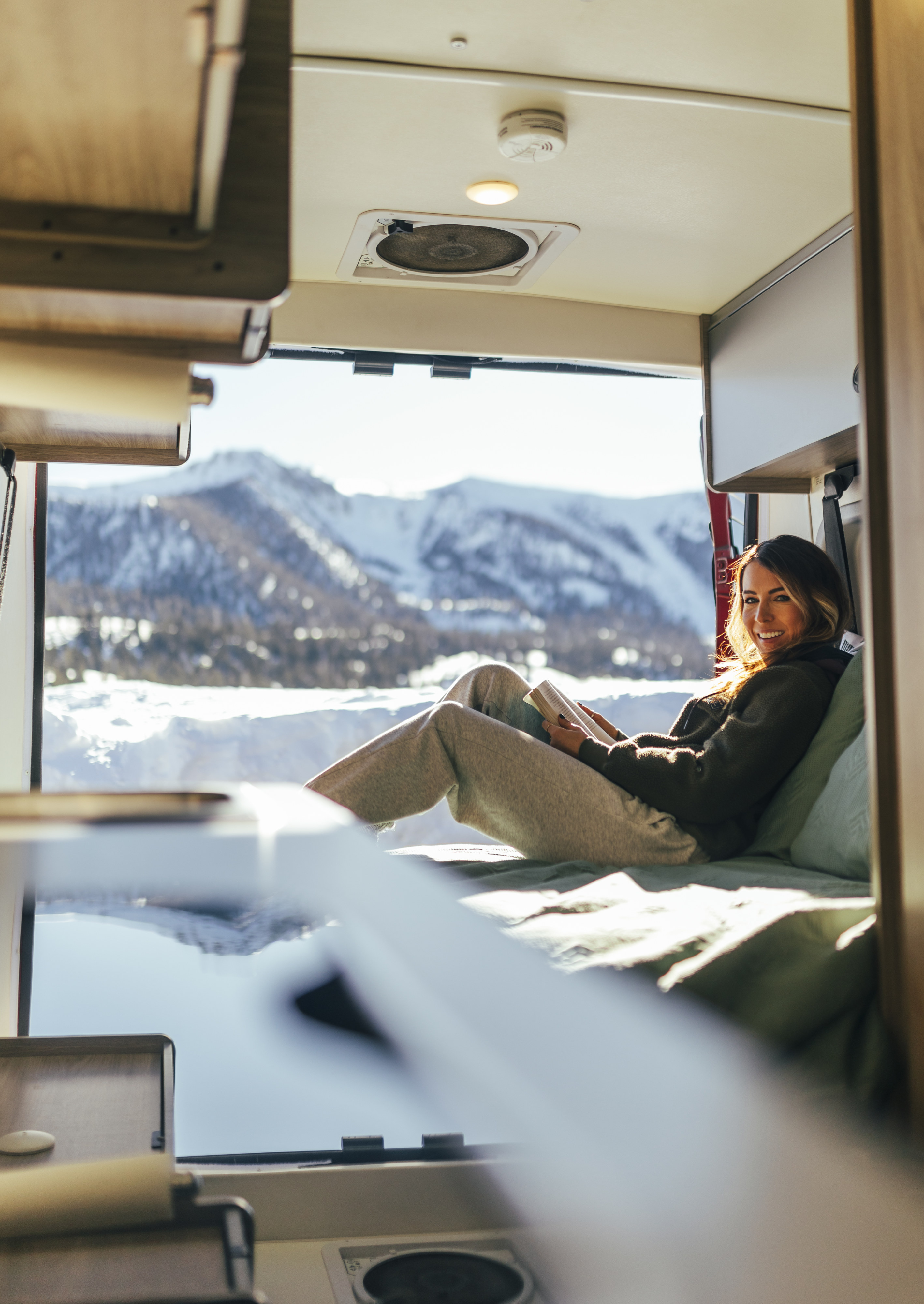 Smiling woman relaxing in a cozy RV rental, reading a book with a stunning view of snowy mountains.. Solis 59P Solis 2022