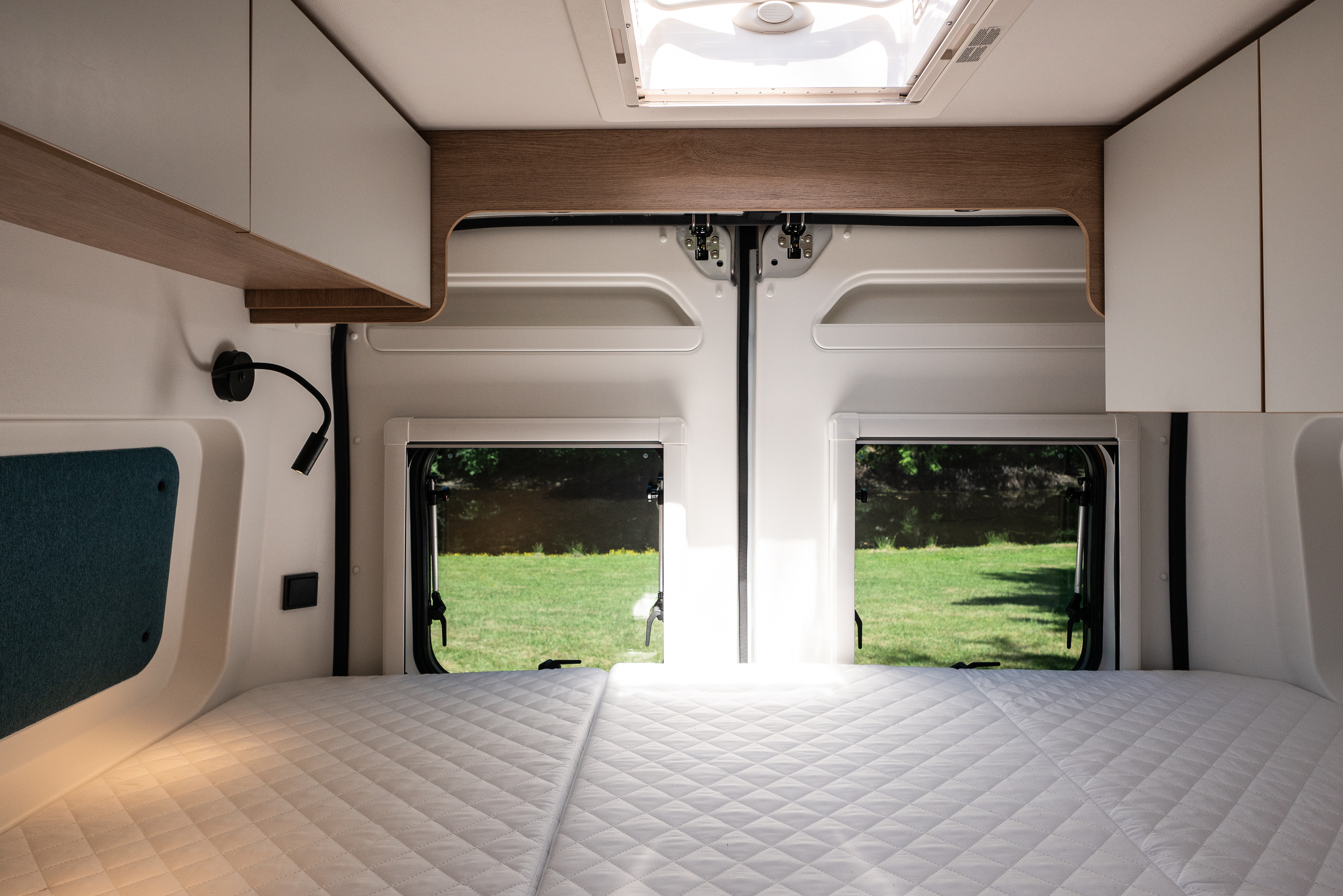 Inside an RV rental: a comfy bed awaits, with scenic views through rear windows and overhead storage.. JOA 60G 2023