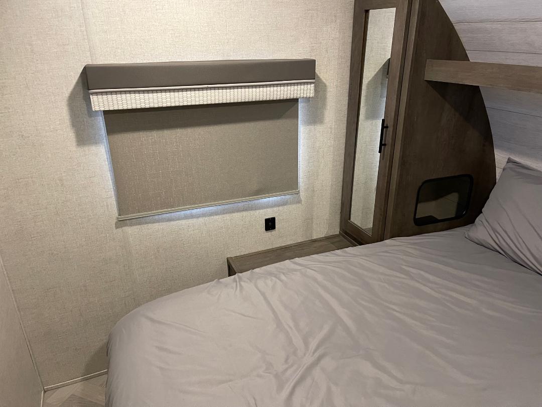 Inside an RV rental bedroom, light gray bedding on a bed, a window with blind, and a wood-toned wardrobe with a mirror.. Forest River Salem Cruise Lite 2022
