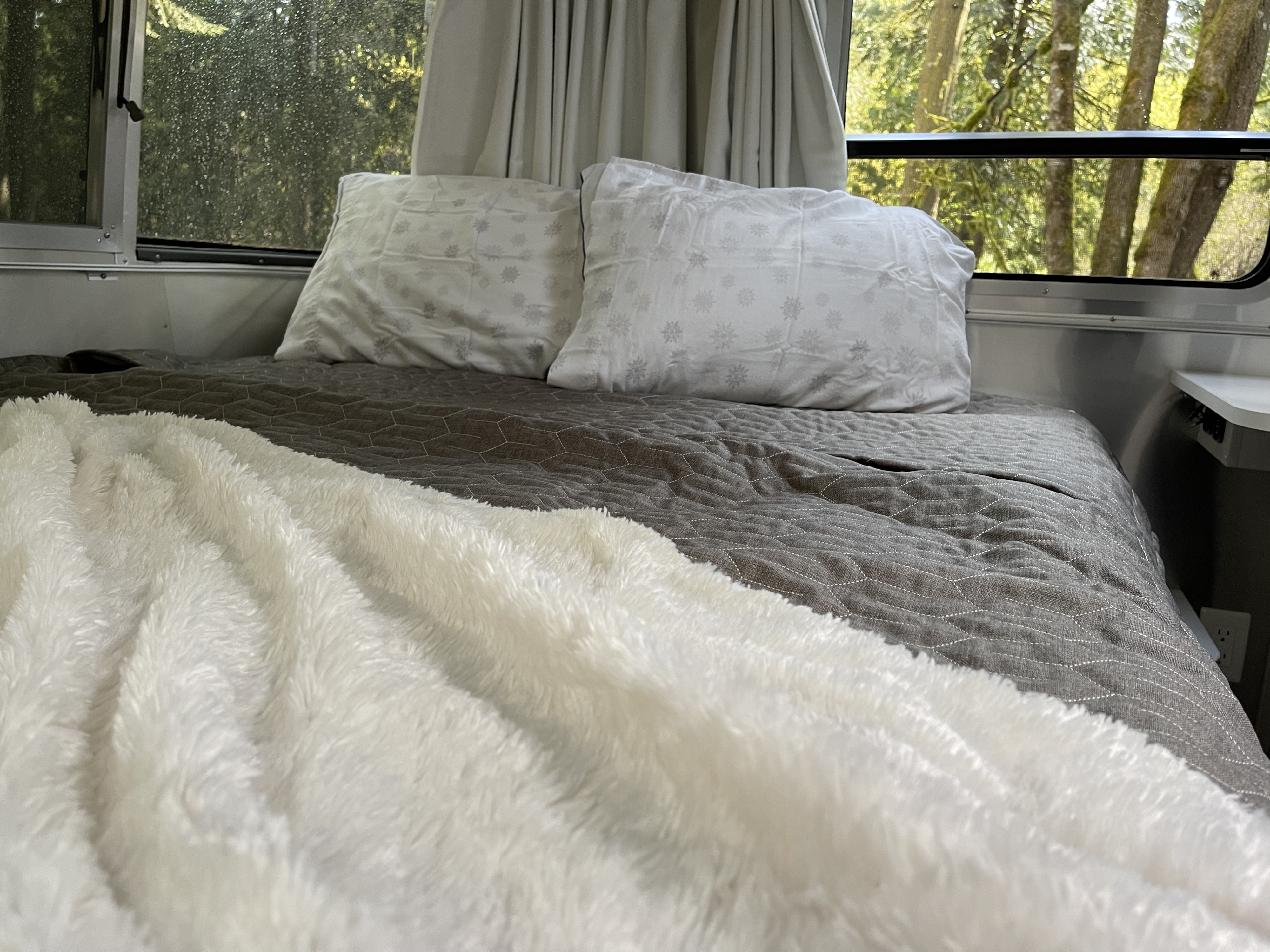 Cozy RV rental bed setup: white pillows, gray geometric quilt, fluffy white blanket. Forest views through the windows.. Airstream Sport 2019
