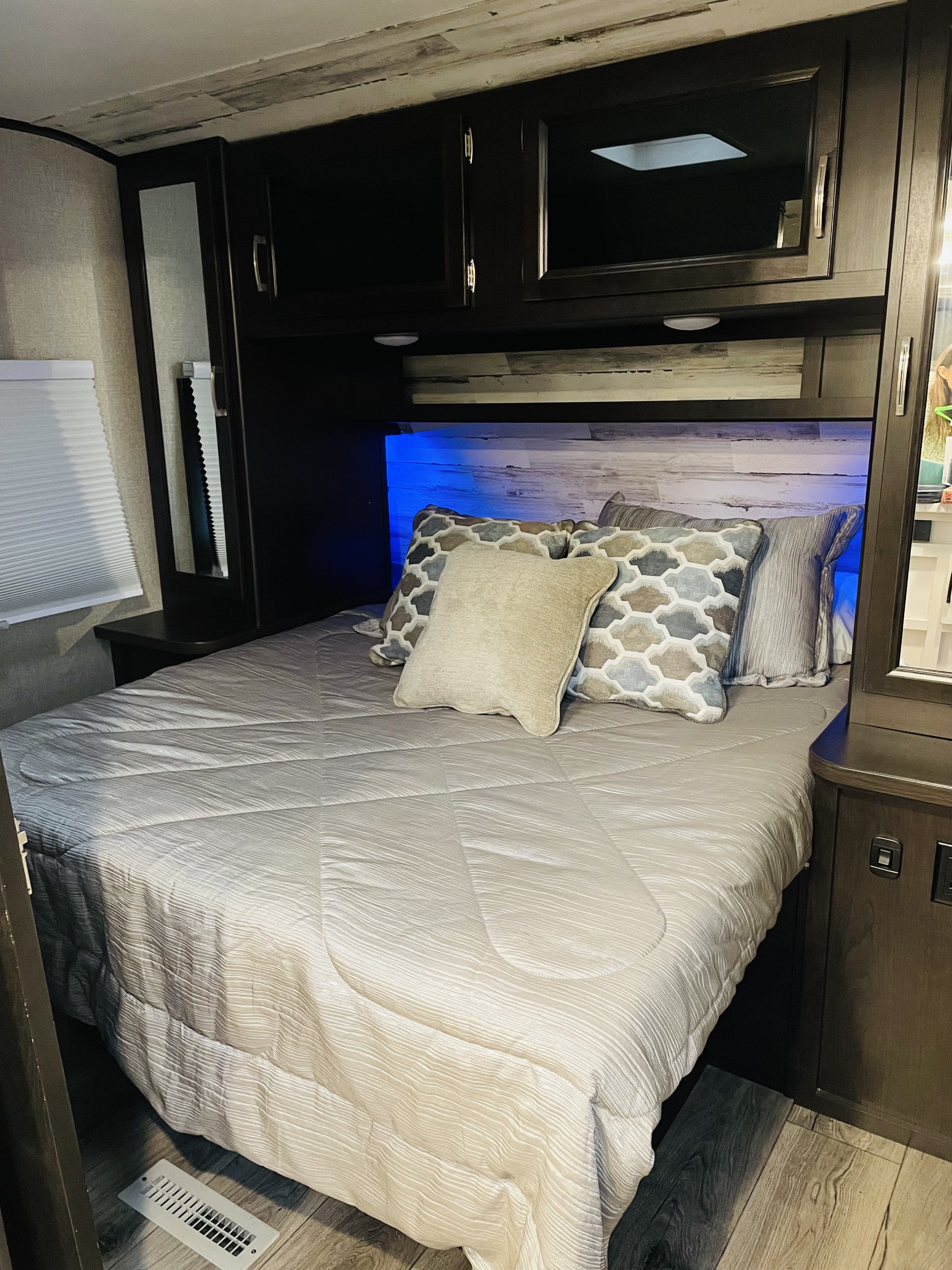 Cozy RV rental bedroom featuring a bed with patterned pillows, dark cabinets, and blue LED headboard lighting.. Jayco Jay Feather 2021