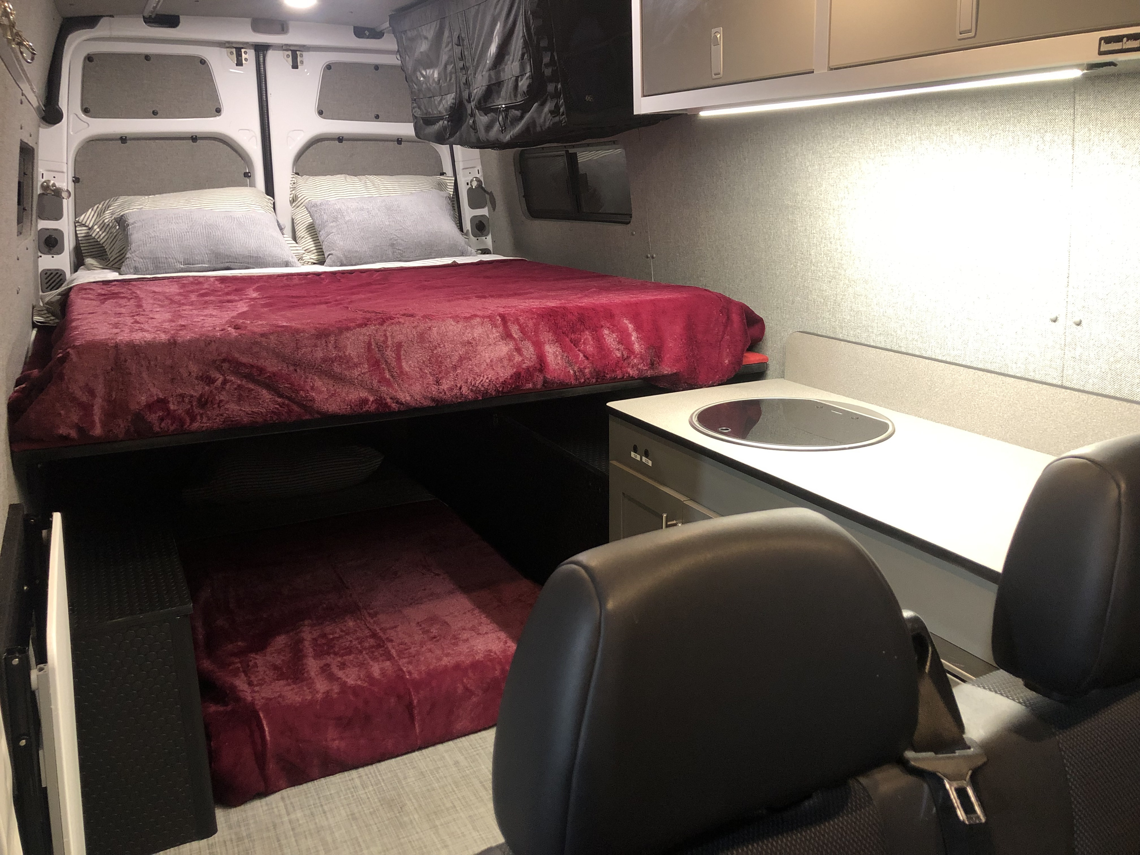Inside an RV rental: a raised bed with red bedding, a lower bunk, and a compact kitchen counter with sink.. Freightliner Sprinter 2012