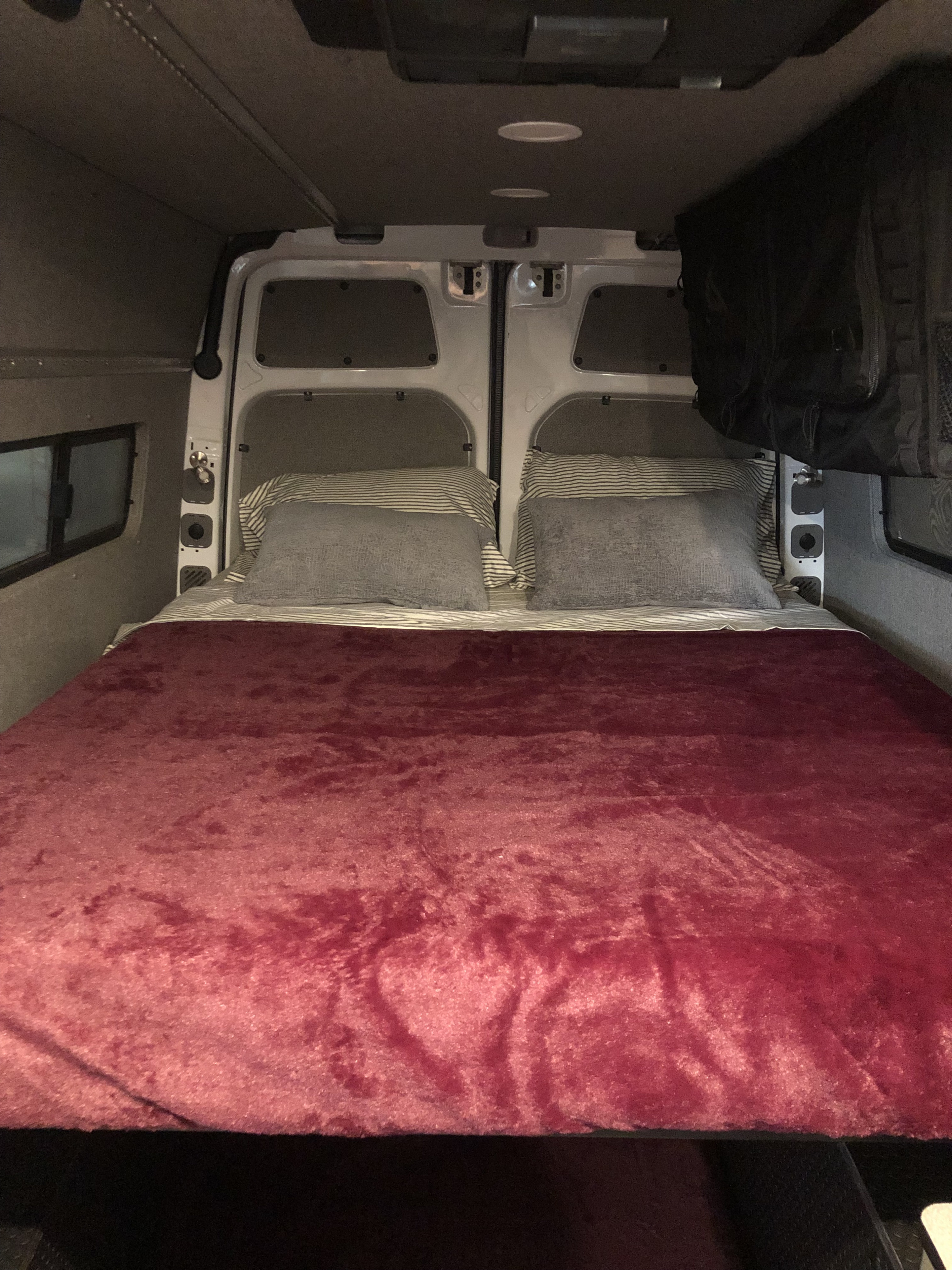 Cozy RV rental van bed with a plush red blanket and pillows, positioned in the vehicle's rear sleeping area.. Freightliner Sprinter 2012