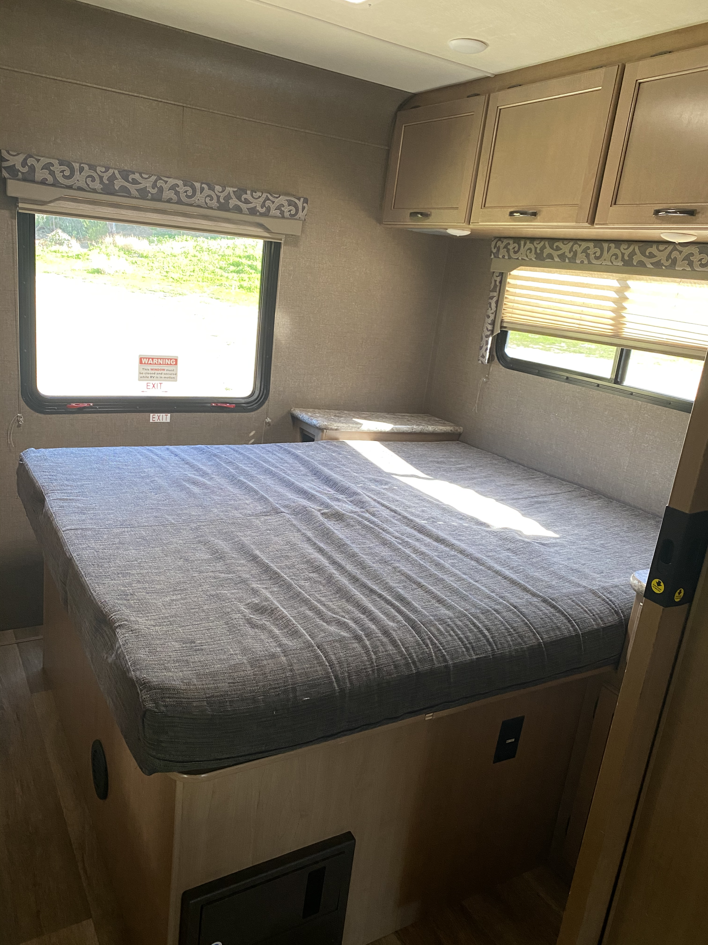 Interior of an RV rental bedroom: bed, overhead cabinets, and bright windows, one with an "EXIT" warning.. Thor Four Winds 2017