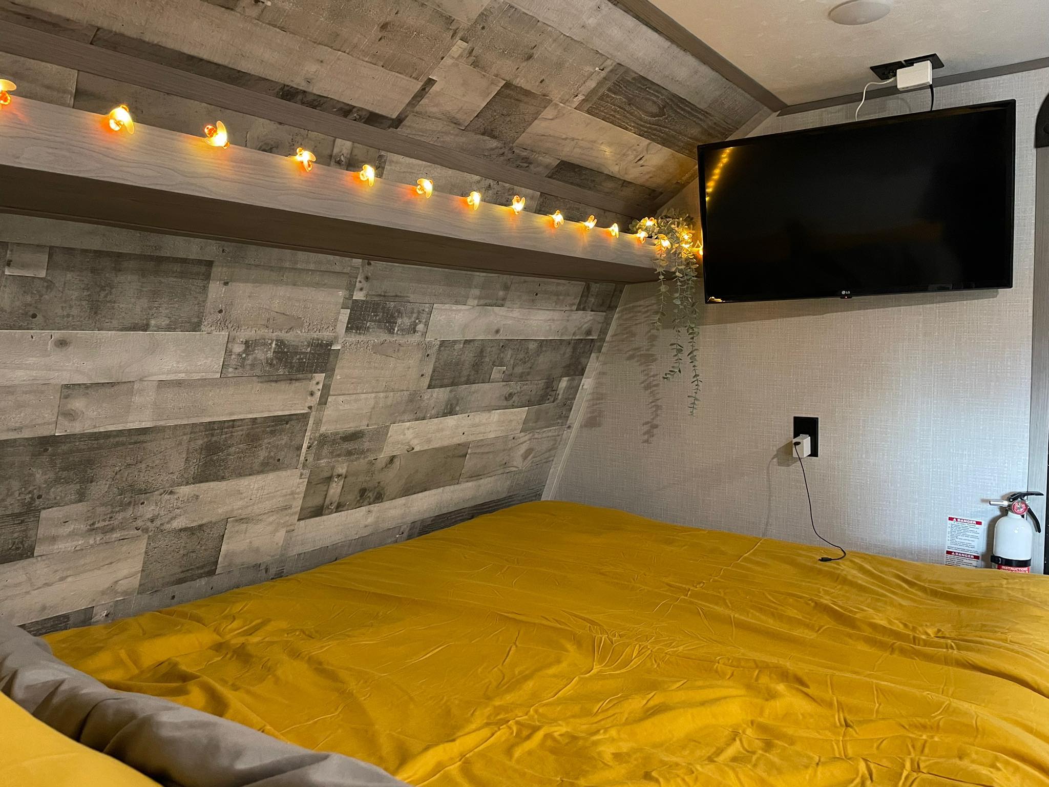 Cozy RV rental bedroom featuring a yellow bed, string lights, mounted TV, and wood-look walls. Fire extinguisher included.. Dutchmen RV Coleman Lantern LT Series 17B 2023