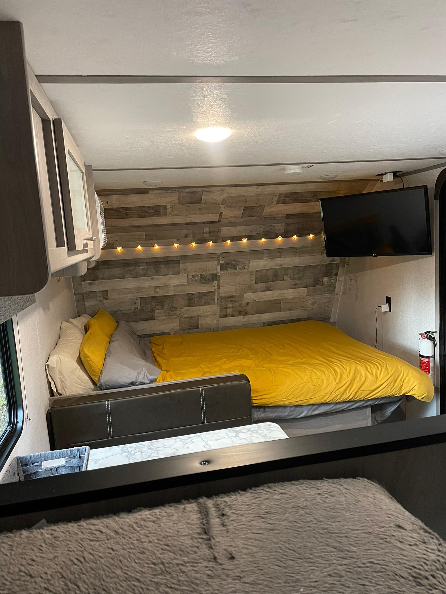 Cozy RV rental interior featuring a bright yellow bed, wood-paneled wall, string lights, and mounted TV. Perfect for glamping adventures.. Dutchmen RV Coleman Lantern LT Series 17B 2023