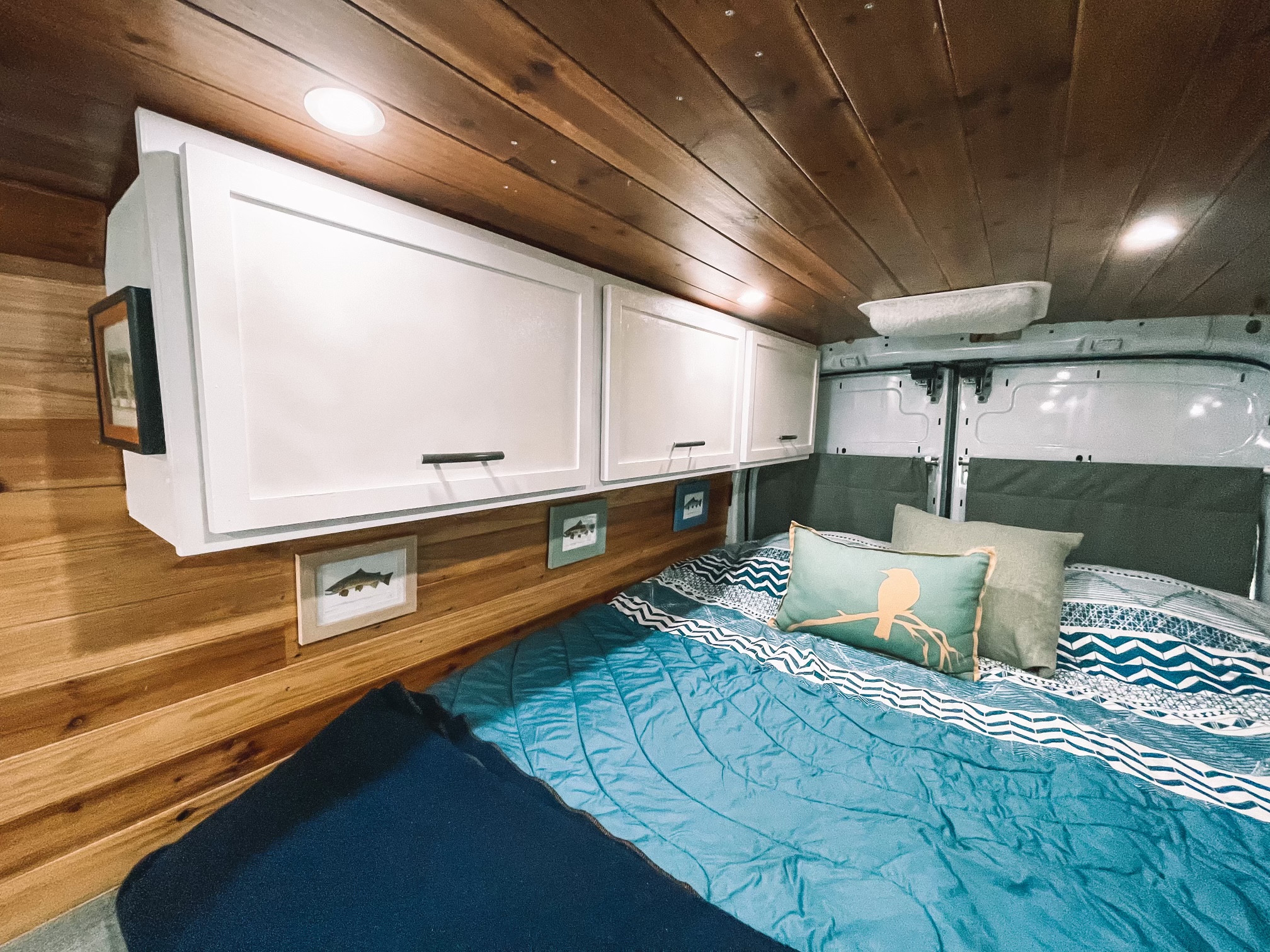 Inside a stylish RV rental: wood-paneled walls & ceiling, white cabinets, & a cozy bed with blue bedding.. Ford Transit Custom 2019