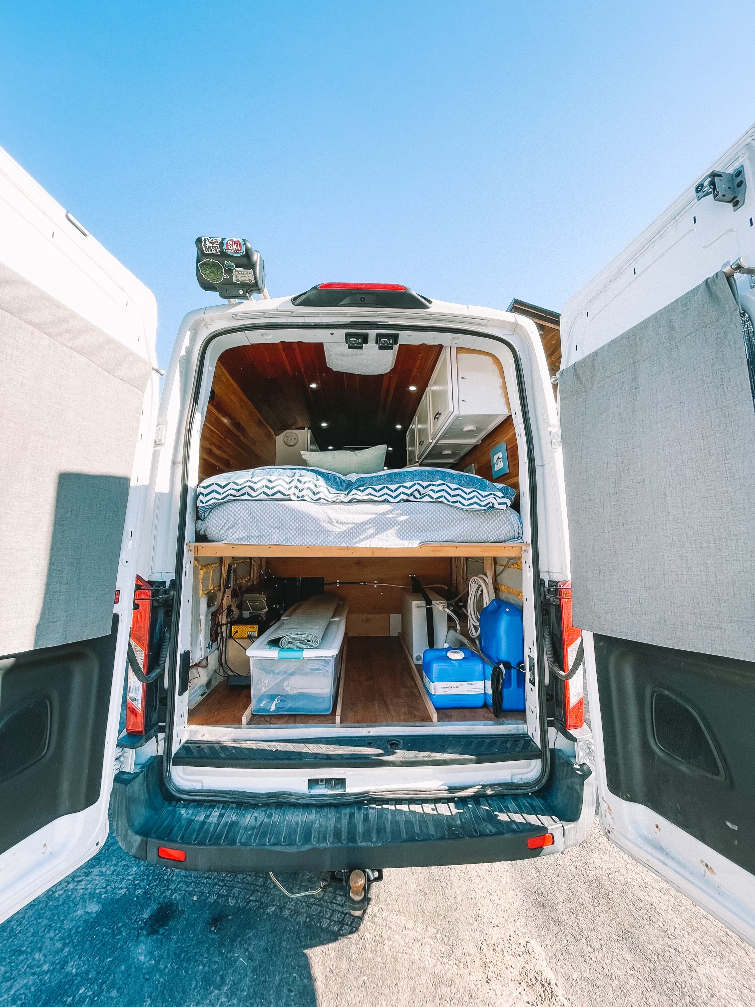 Rear view into a white custom camper van's open interior, revealing a raised bed, organized storage, and utilities. Great for RV rental.. Ford Transit Custom 2019