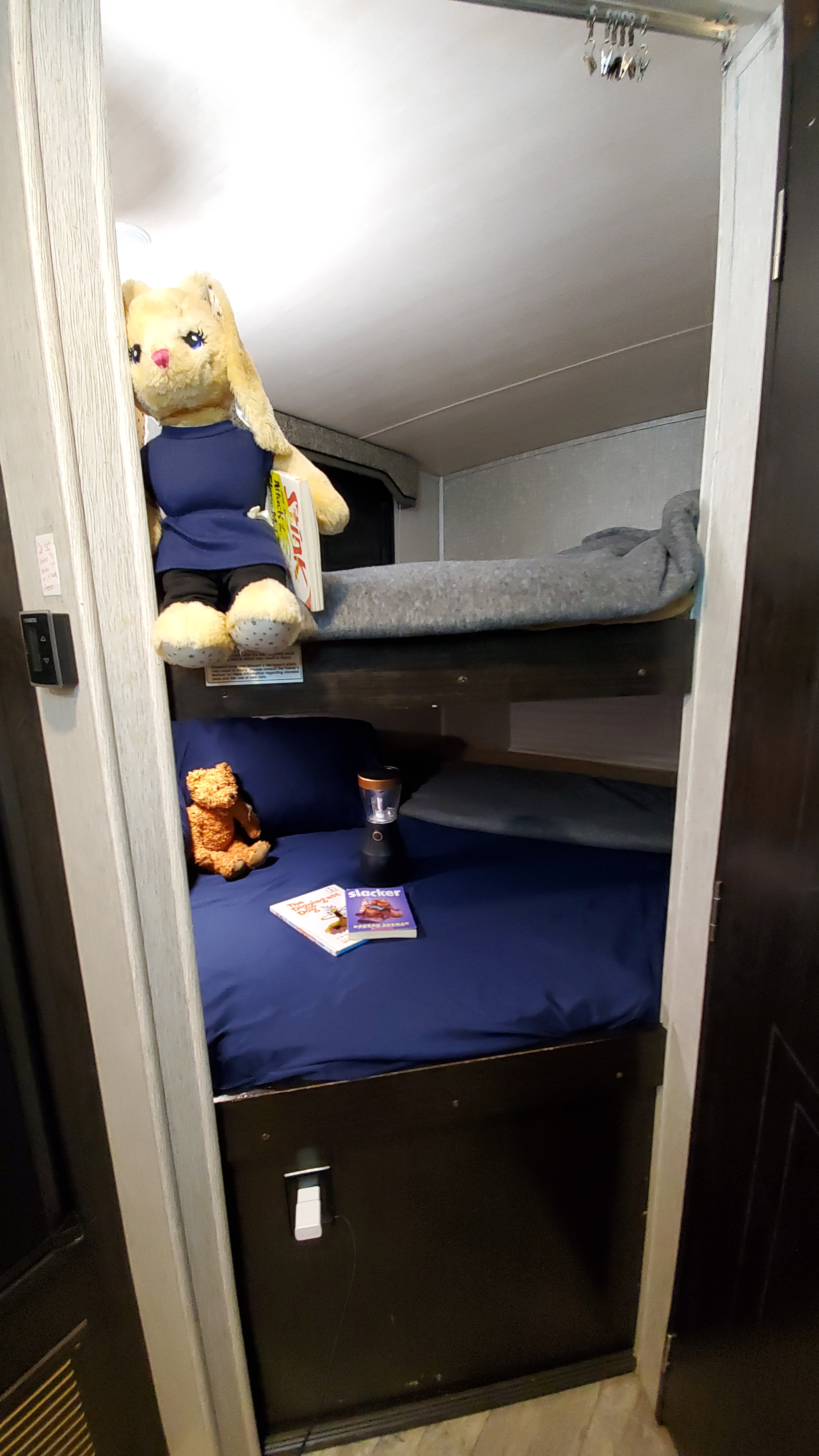 RV rental interior: Stacked bunk beds with blue sheets, a gray blanket, plush toys (rabbit, bear), books, & a lantern. Cozy sleeping area.. Heartland RVs North Trail 2018