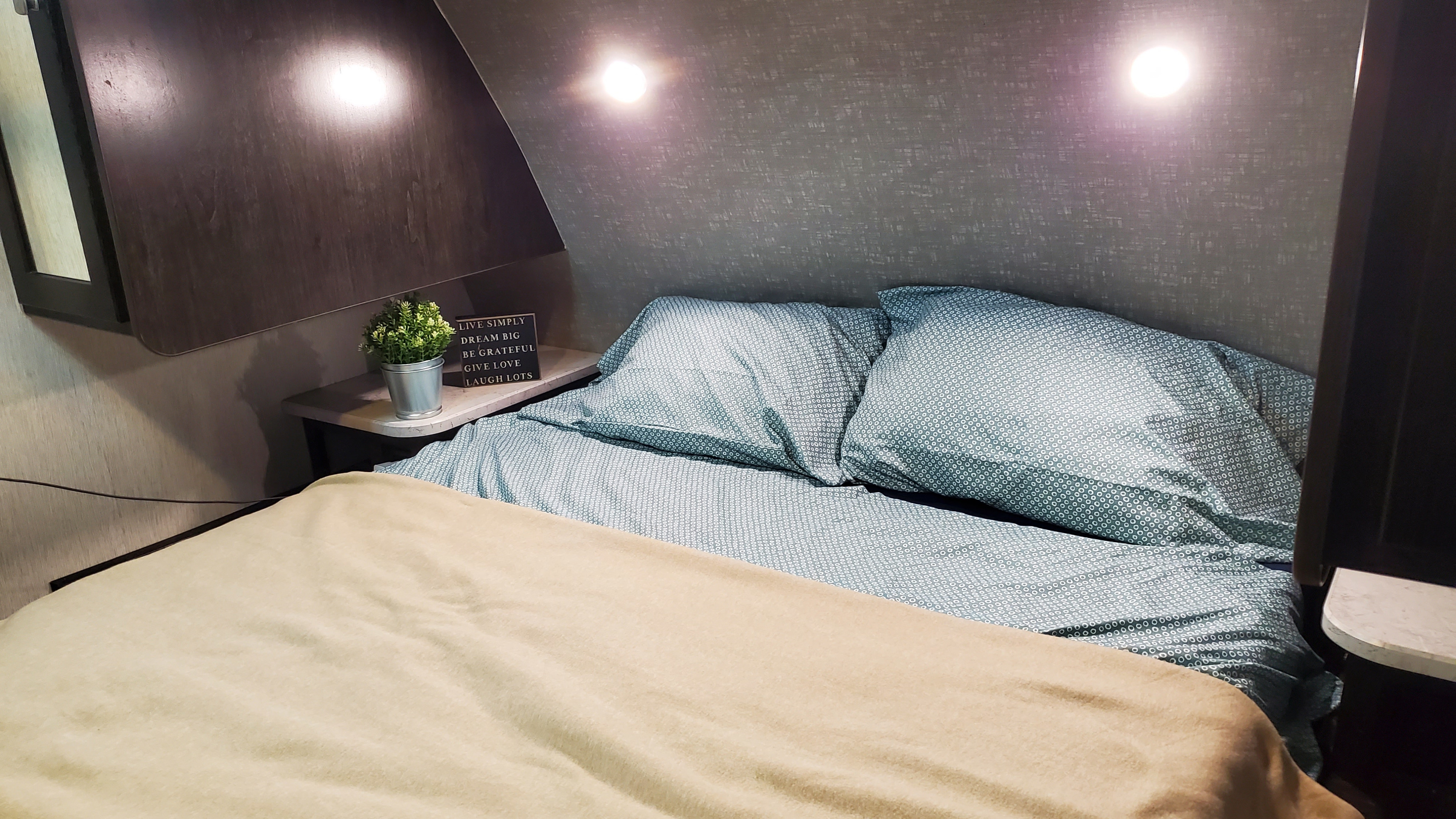 Cozy RV rental bedroom featuring a comfortable bed with patterned sheets, nightstand with plant & sign, and overhead lighting.. Heartland RVs North Trail 2018