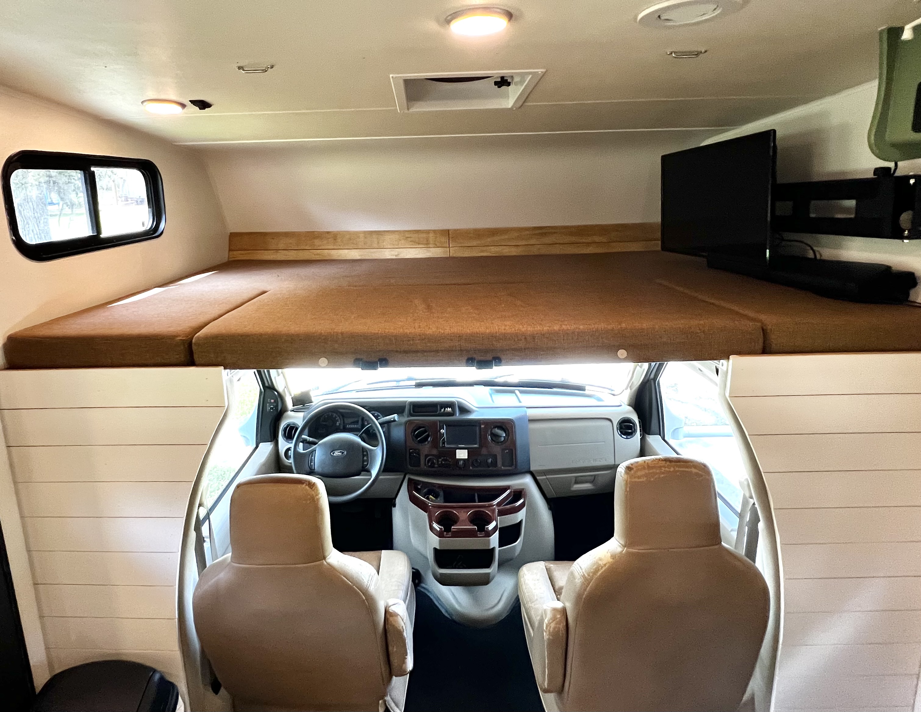 Interior of an RV rental showing a comfortable cab-over bunk with window & TV, overlooking the Ford driver's cab.. Forest River Coachmen Leprechaun 2016