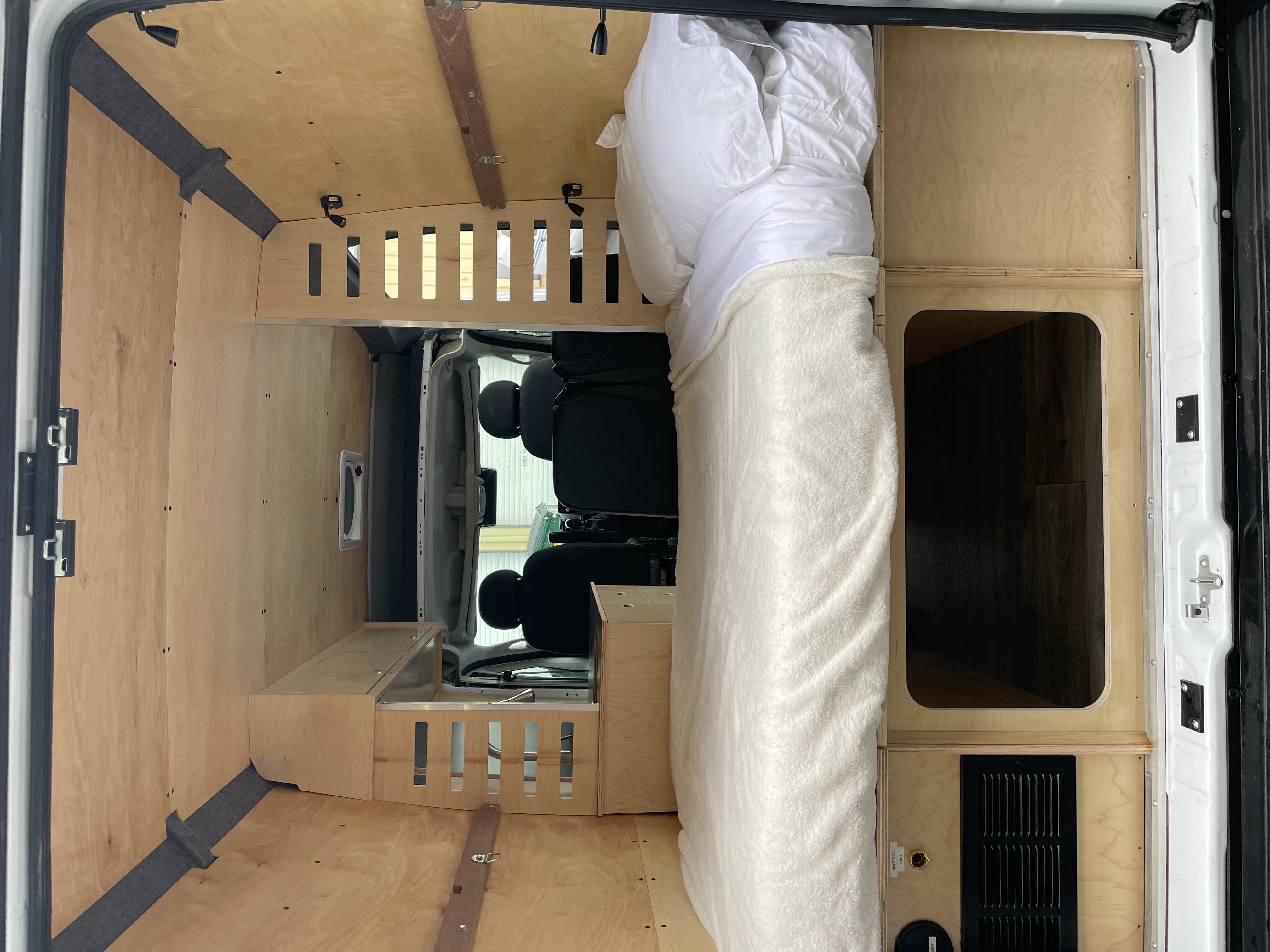Wood-paneled RV rental interior with a cozy bed, integrated storage, and views into the front cabin.. RAM ProMaster 2023