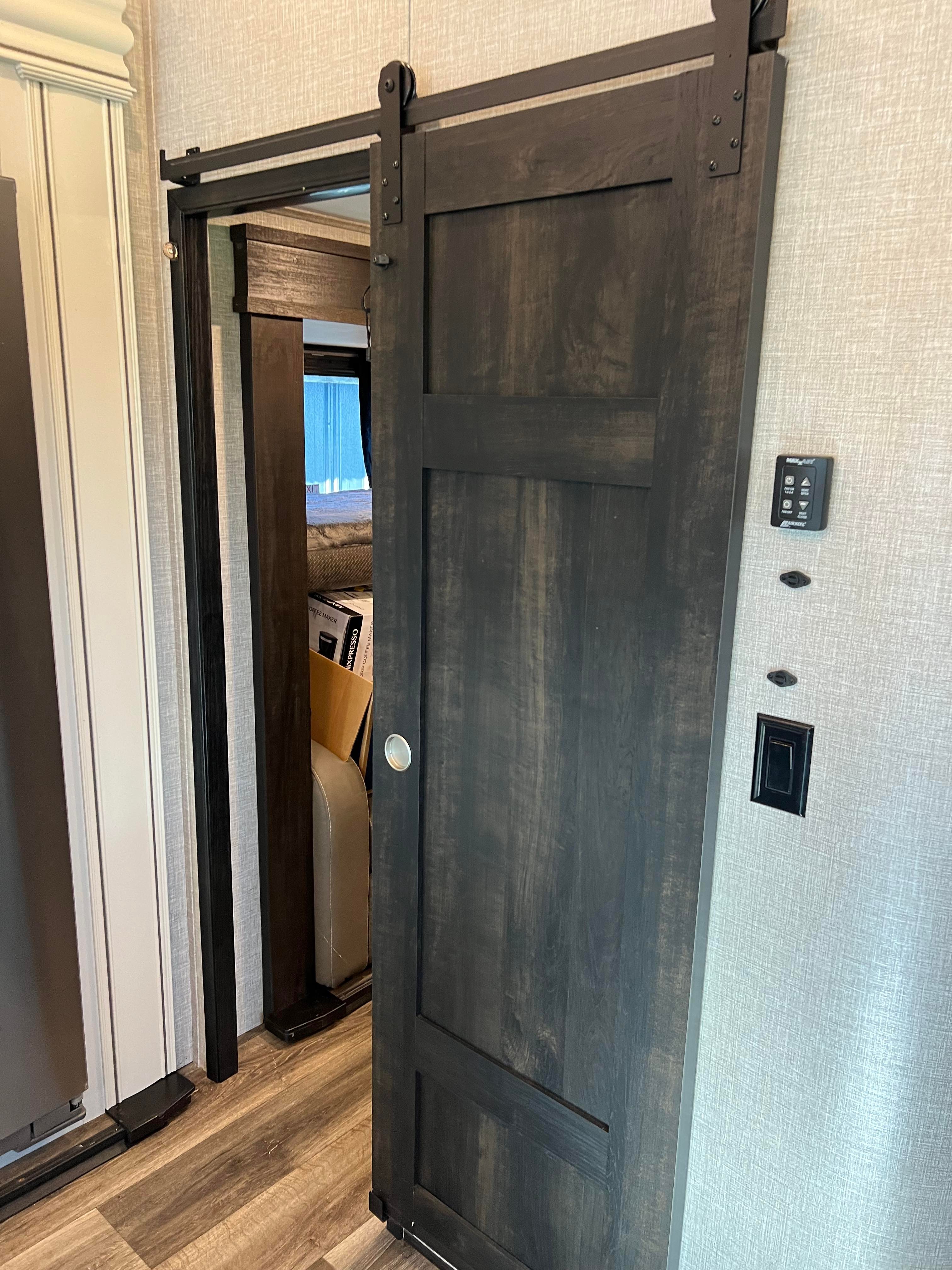 Inside a stylish RV rental: a dark wood-grain barn door slides open to a bedroom. Light walls, modern controls, and wood flooring.. Jayco North Point 2022