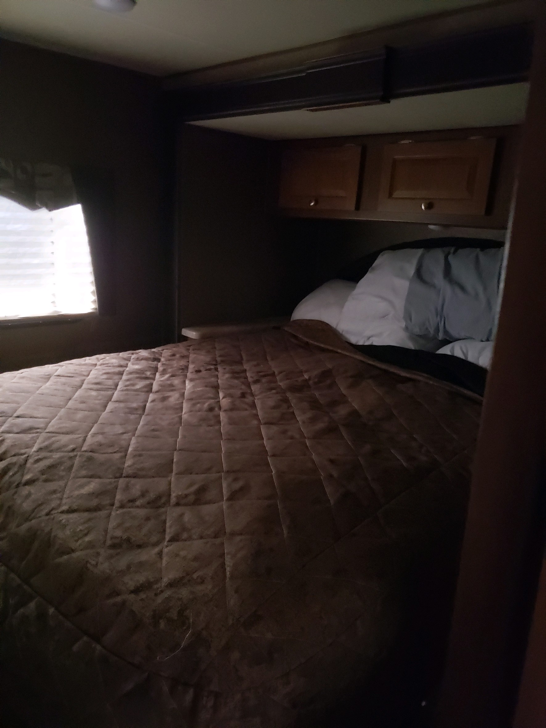 Compact RV rental bedroom featuring a brown quilted bed, storage cabinets, and window with blinds.. Coachman Leprechaun 2017