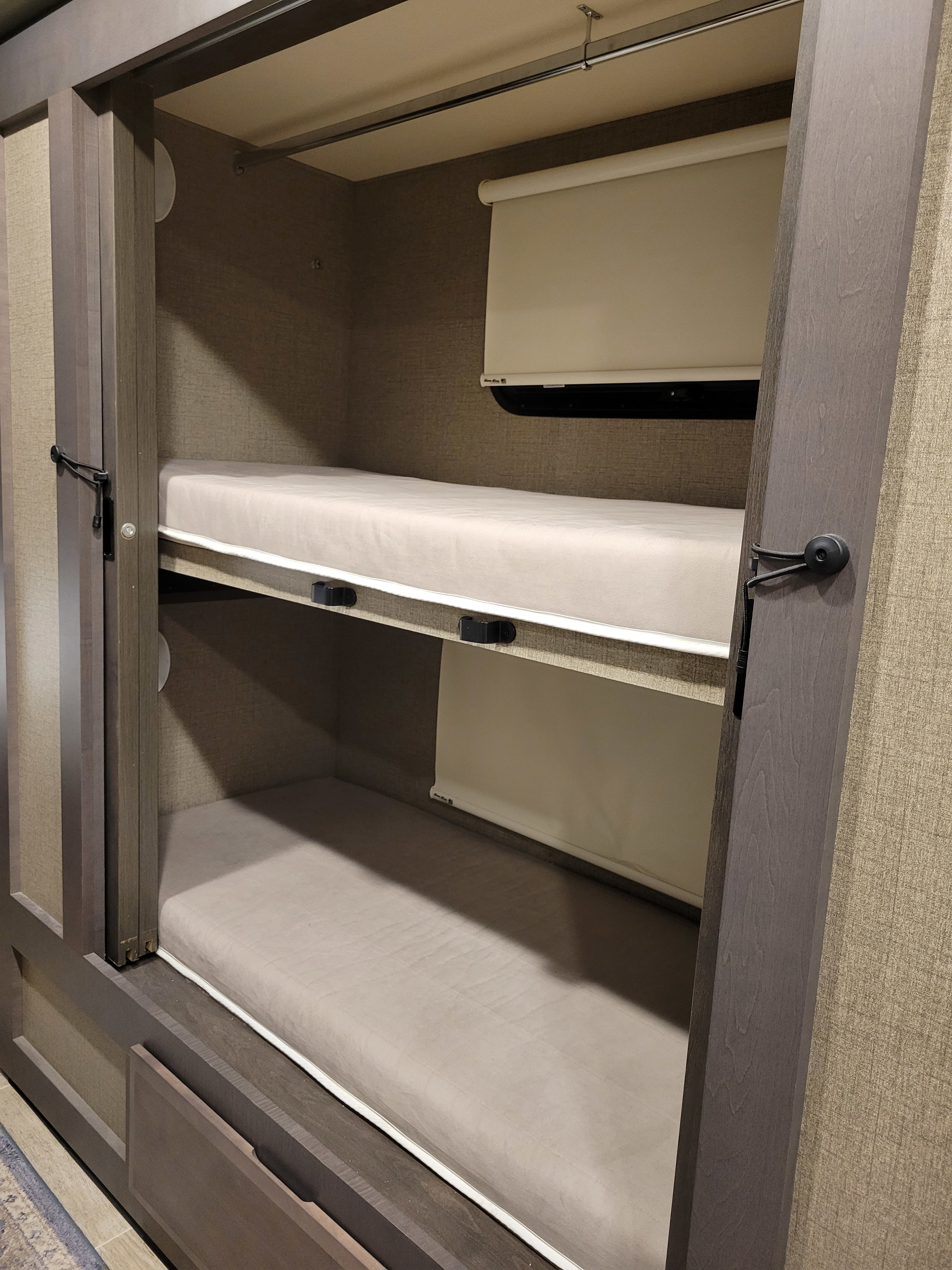 Interior view of an RV rental showing two neatly made bunk beds with mattresses, integrated roller blinds, and a top clothing rod.. Thor Motor Coach Omni 2022