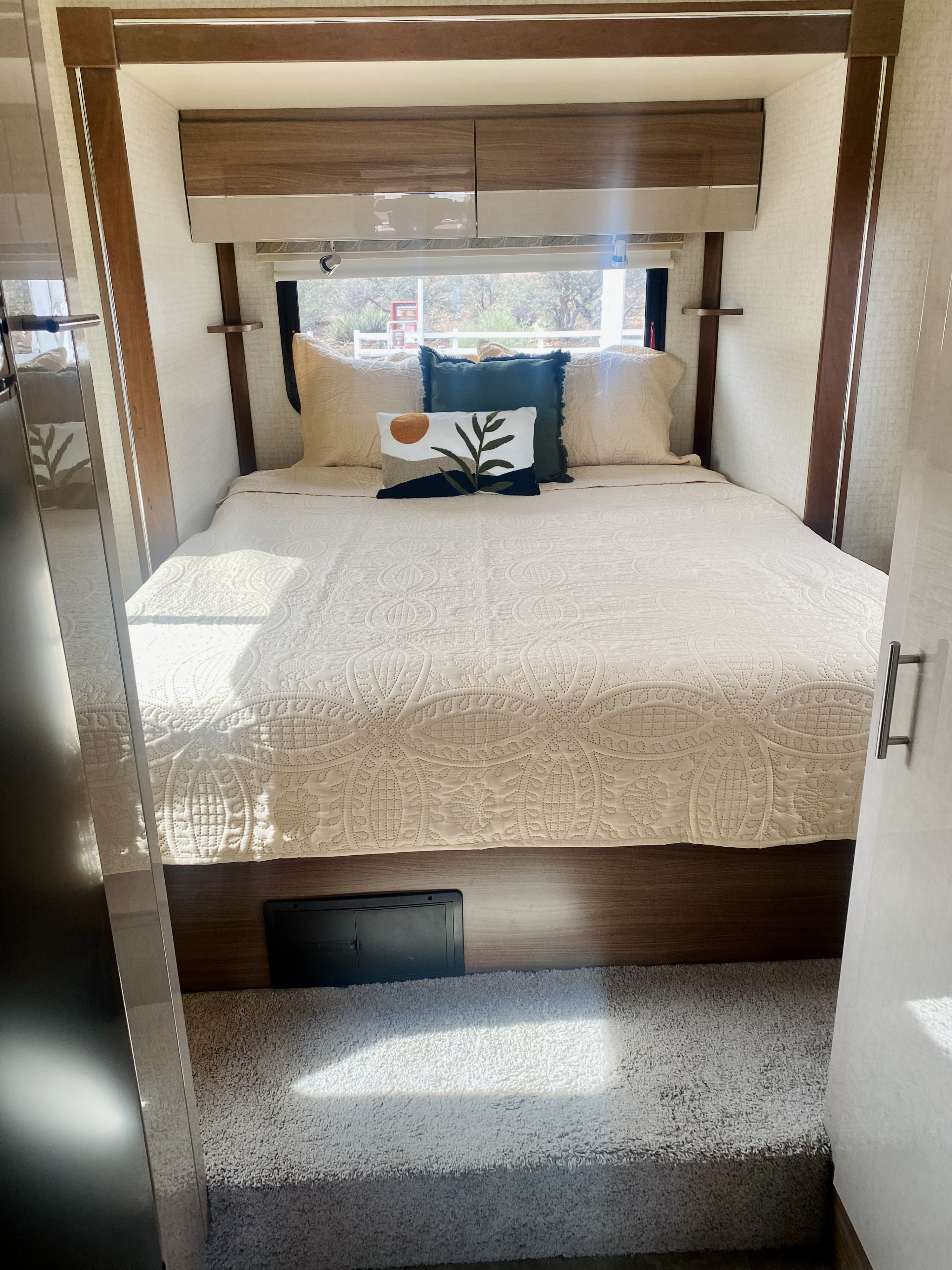 Cozy RV rental bedroom with a quilted bed, decorative pillows, overhead storage, and a sunny window view.. Winnebago View 2016