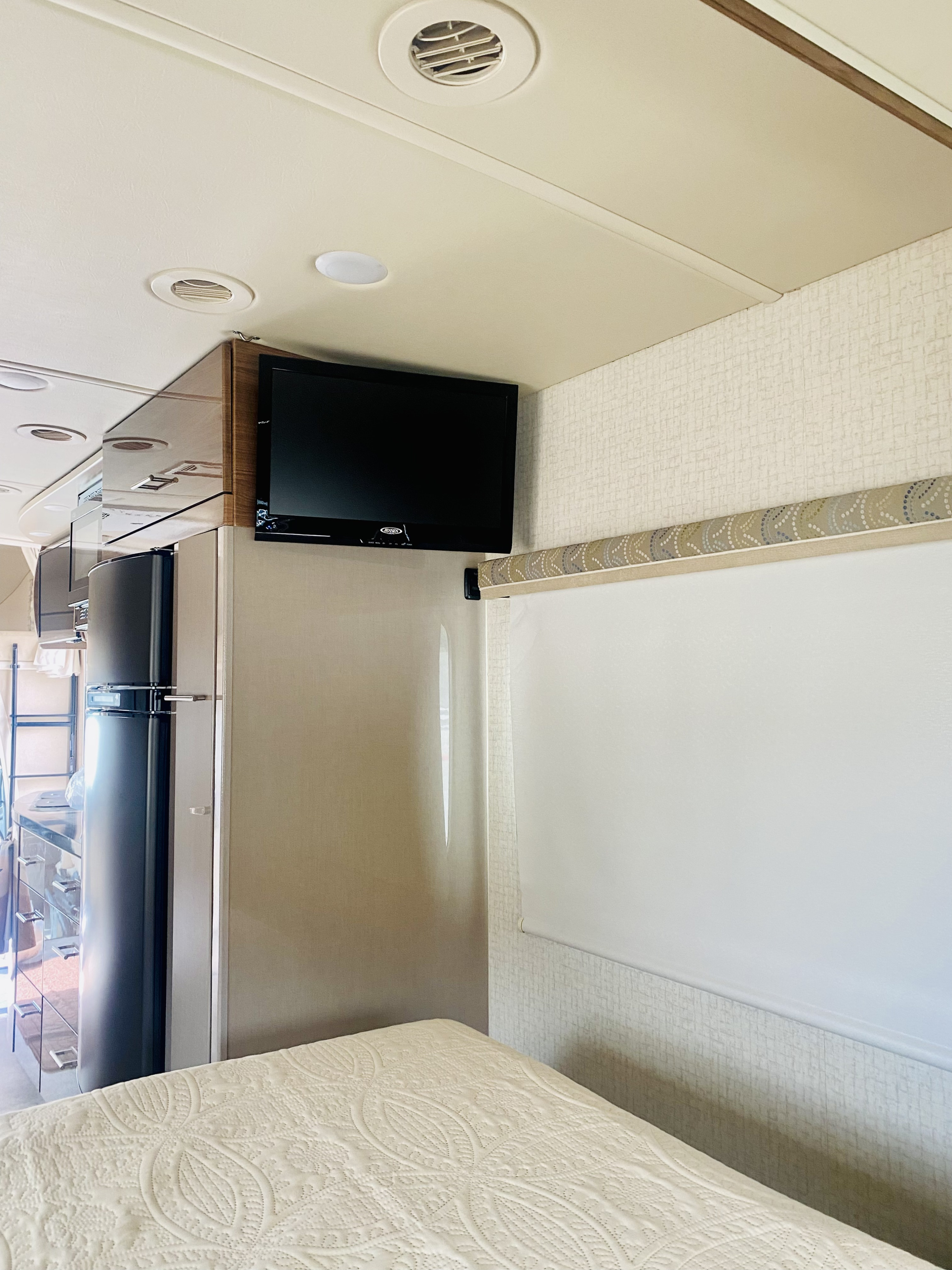 Inside an RV rental: A compact bedroom area with a patterned bed, mounted TV, built-in fridge, and window blind.. Winnebago View 2016