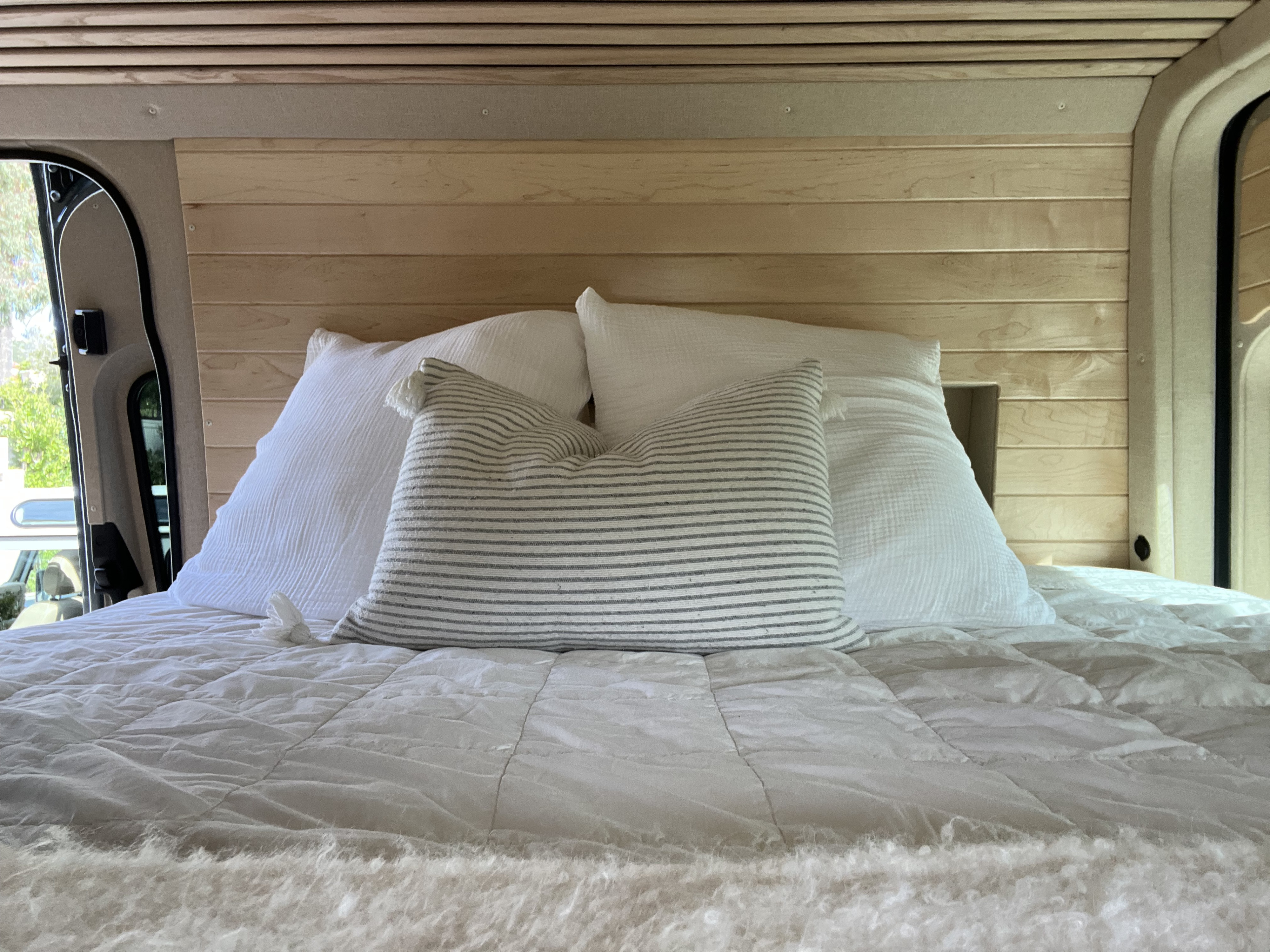 Inside a comfy RV rental: a bed with white & striped pillows, quilted blanket, wood walls, and a window view.. Mercedes Sprinter 2022
