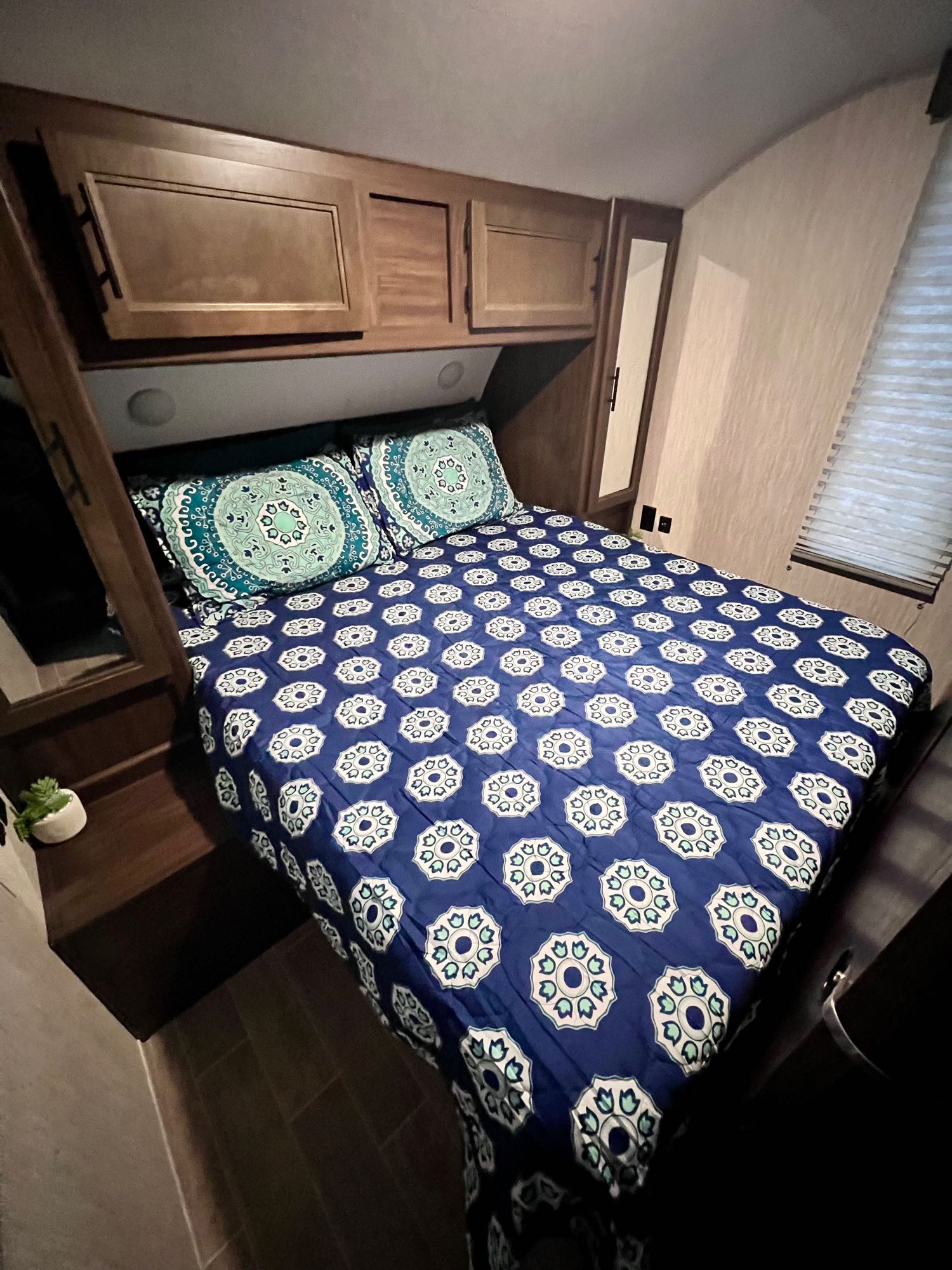 RV rental bedroom with vibrant blue and green patterned bedspread, overhead wooden cabinets, and a side window.. Forest River Cherokee Grey Wolf 2021