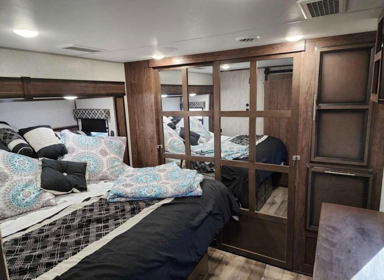 Inside an RV rental bedroom, showing a bed with blue/black patterned bedding and a mirrored sliding closet door for storage.. KZ Durango 2021