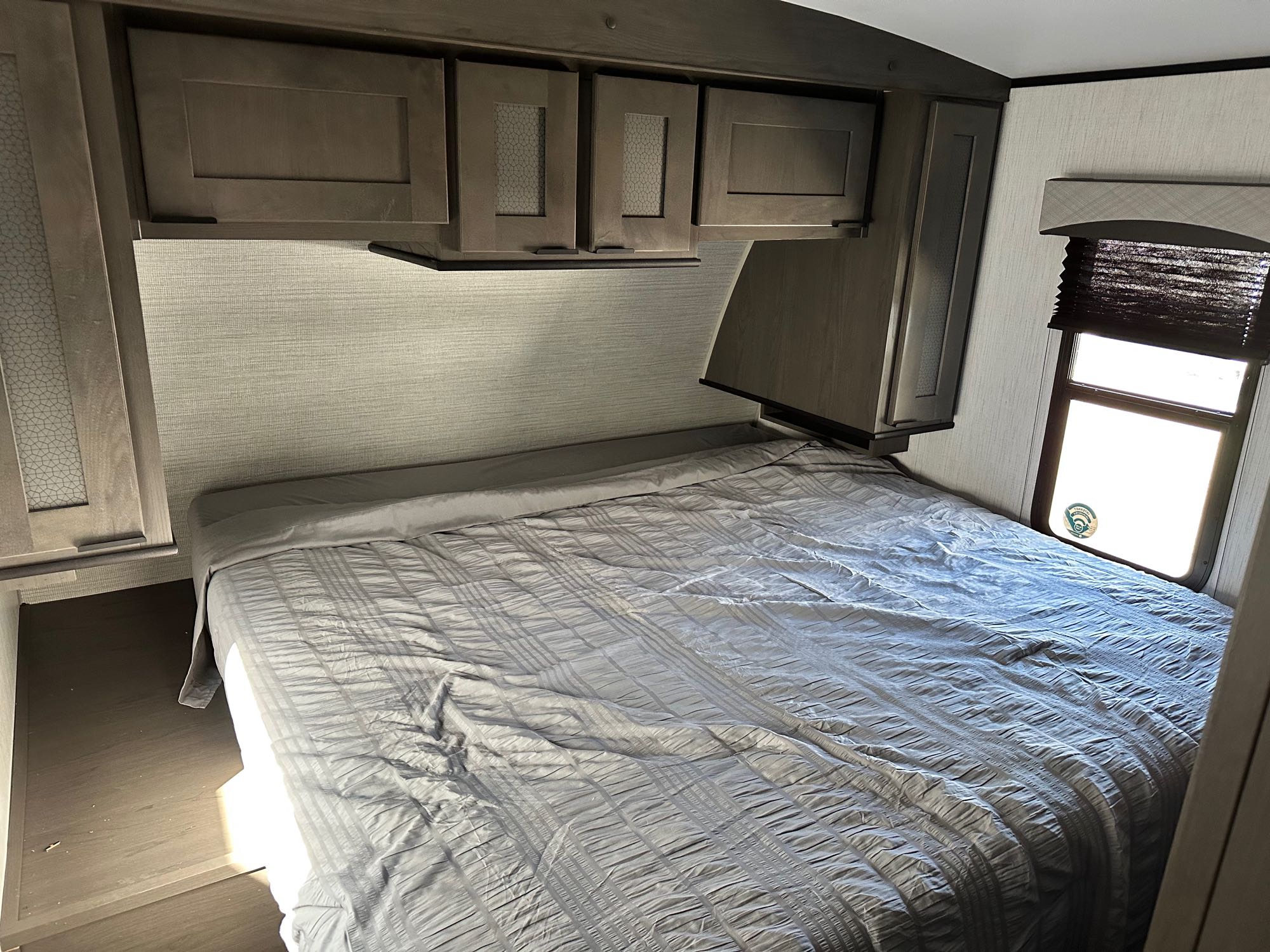 View inside an RV rental bedroom showing a gray bed, overhead cabinets, and a window with blinds.. Cruiser RV Embrace Ultra Lite 2022