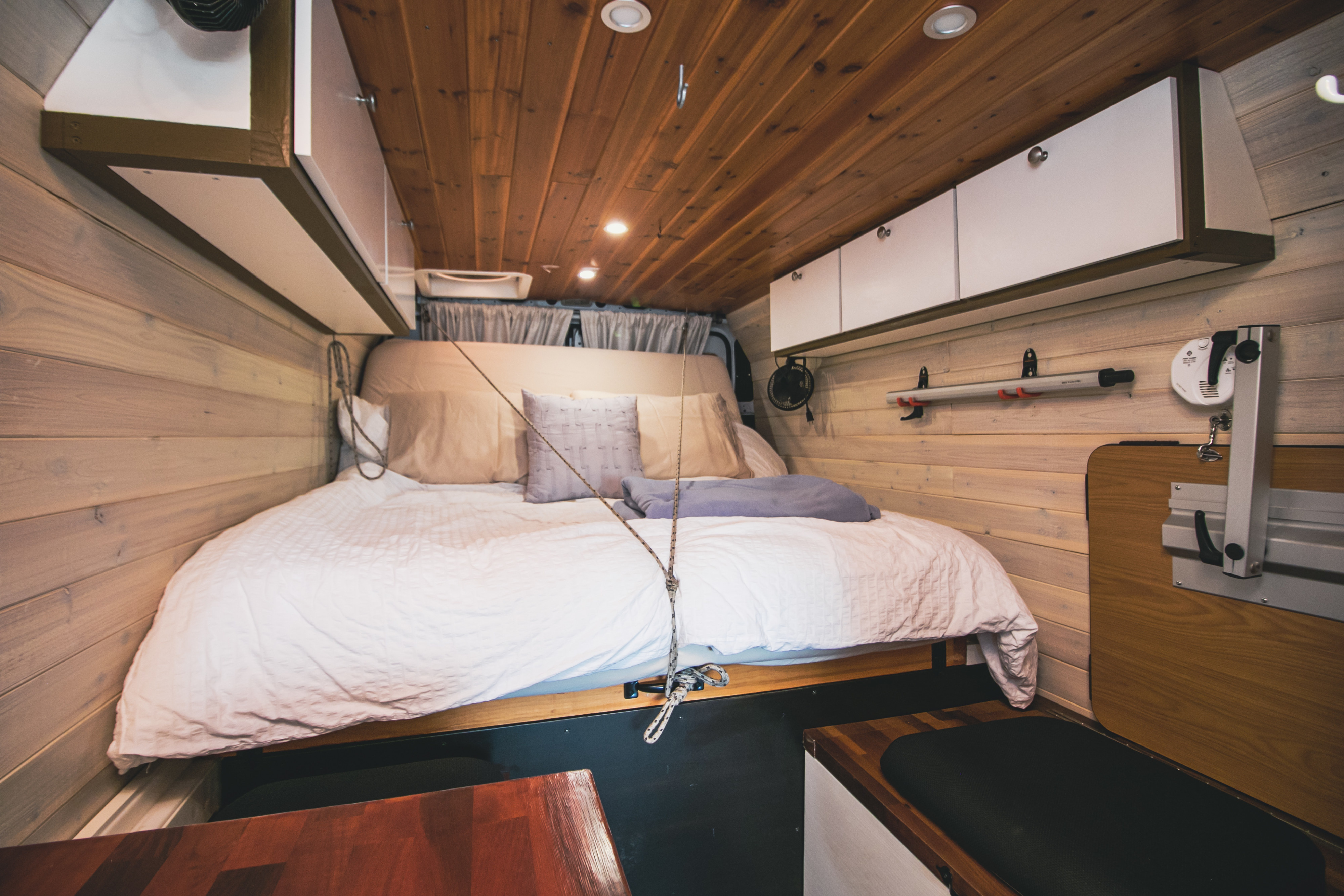 Interior of an rv rental camper van, featuring a cozy bed, wooden walls & ceiling, overhead storage, and a table system.. RAM Promaster 3500 2020